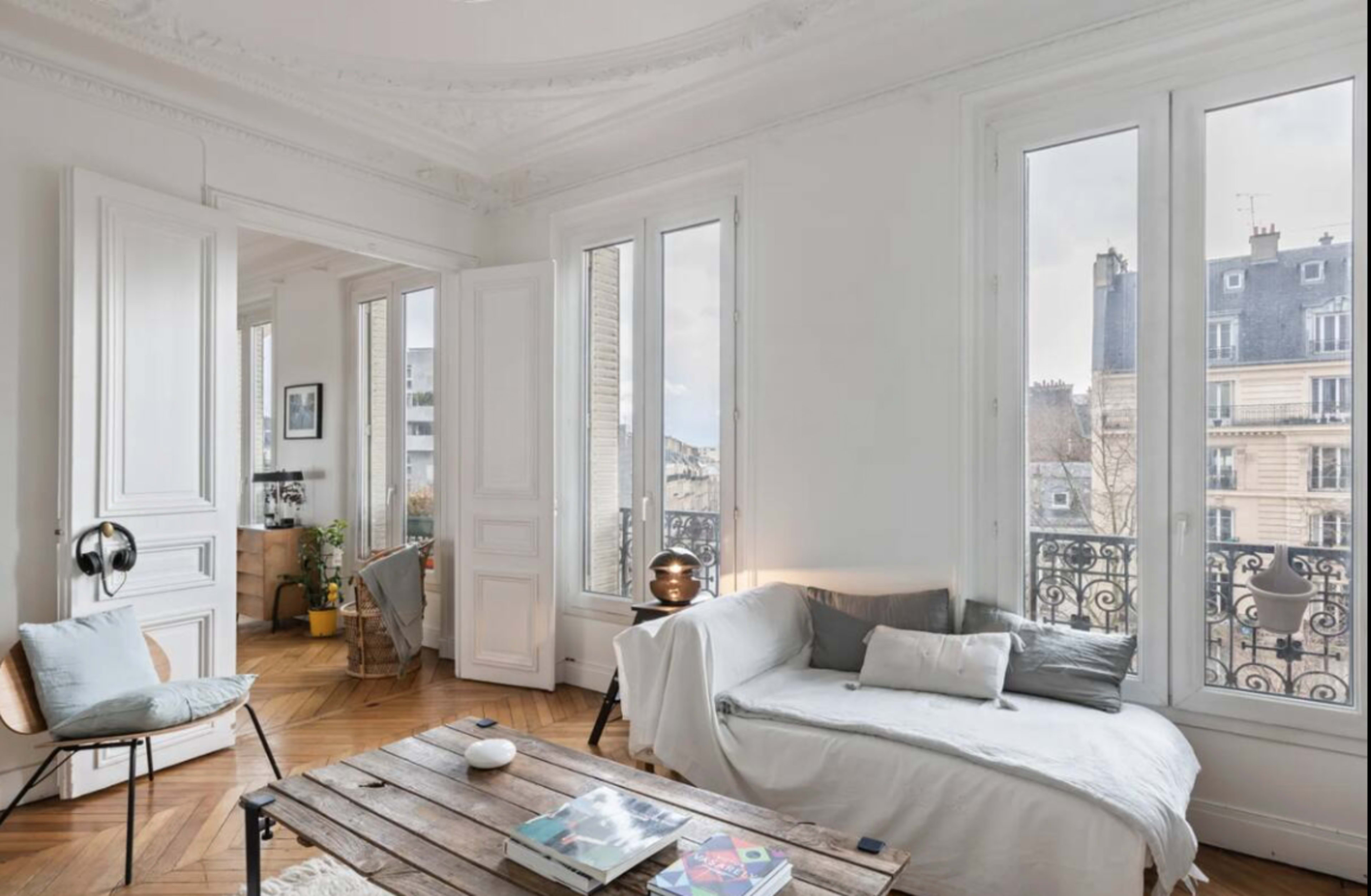 The image shows a bright, contemporary living room with large windows, a wooden coffee table, and a light-colored sofa.