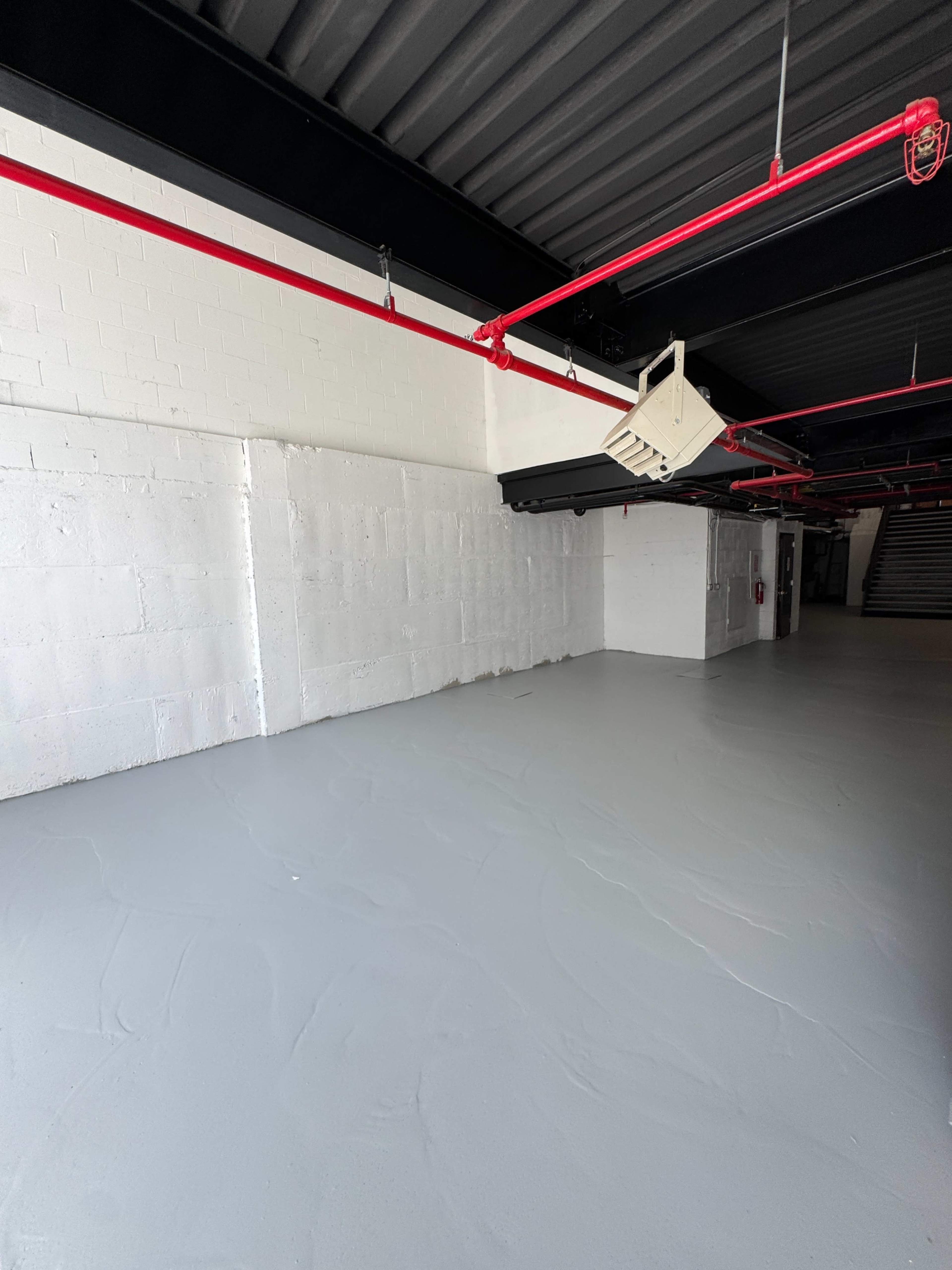 The image shows a spacious, empty room with a smooth gray floor and red pipes running along the ceiling.