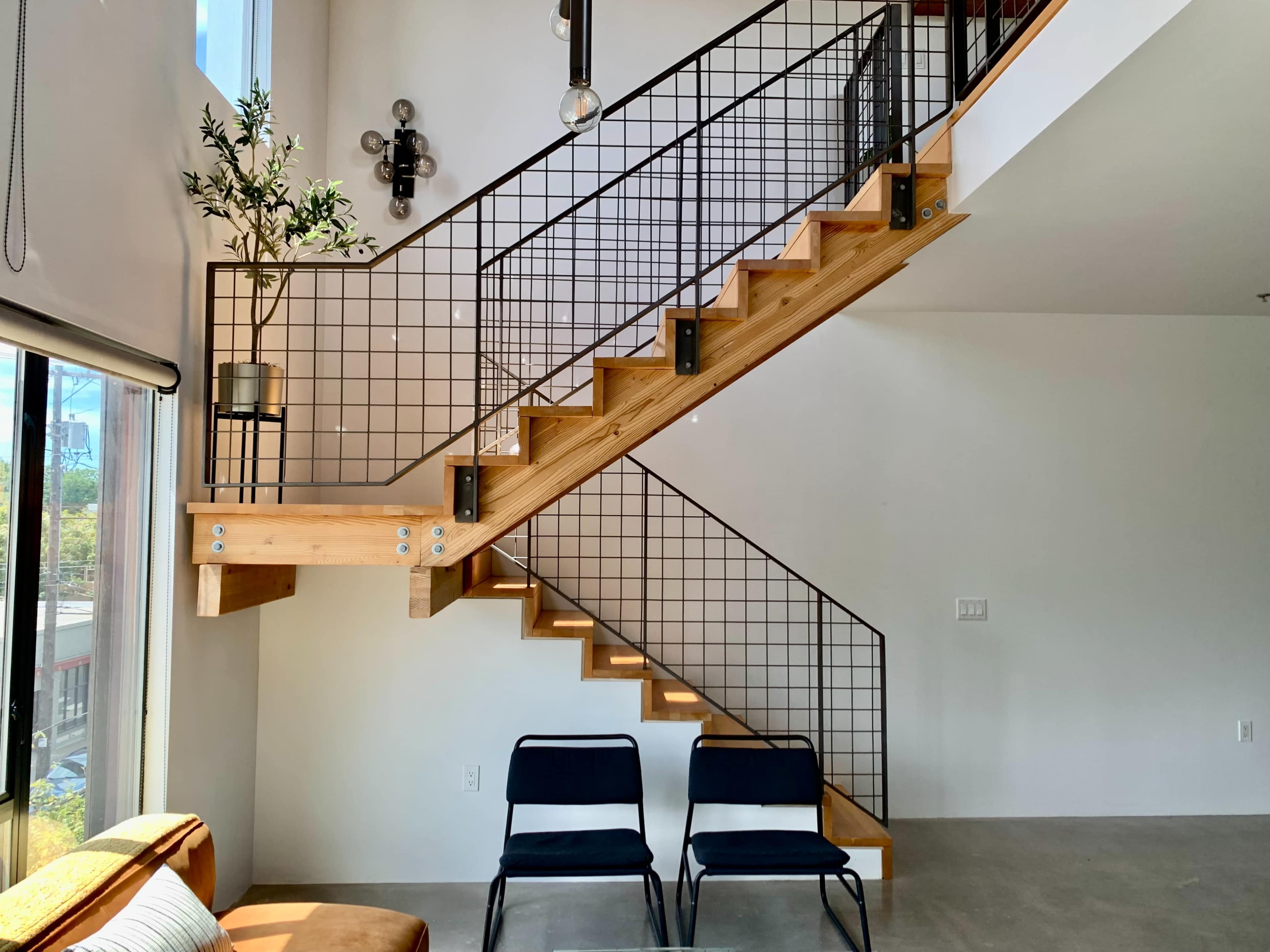A modern staircase made of wood with a metal railing, situated next to large windows and two black chairs on a concrete floor.