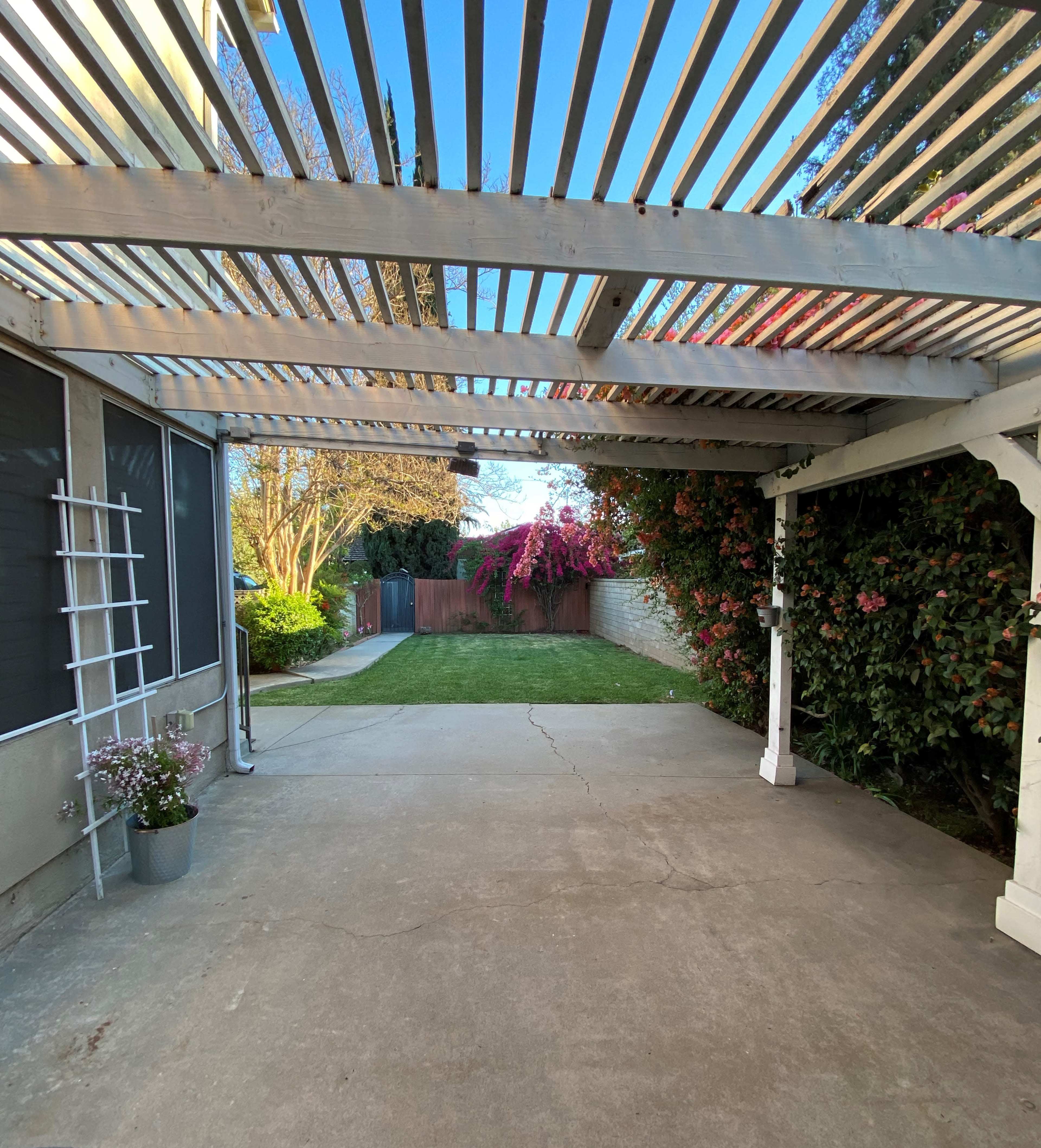 The image shows a patio area with a slatted overhead structure, concrete flooring, and green grass beyond, bordered by flowering plants and a wooden fence.