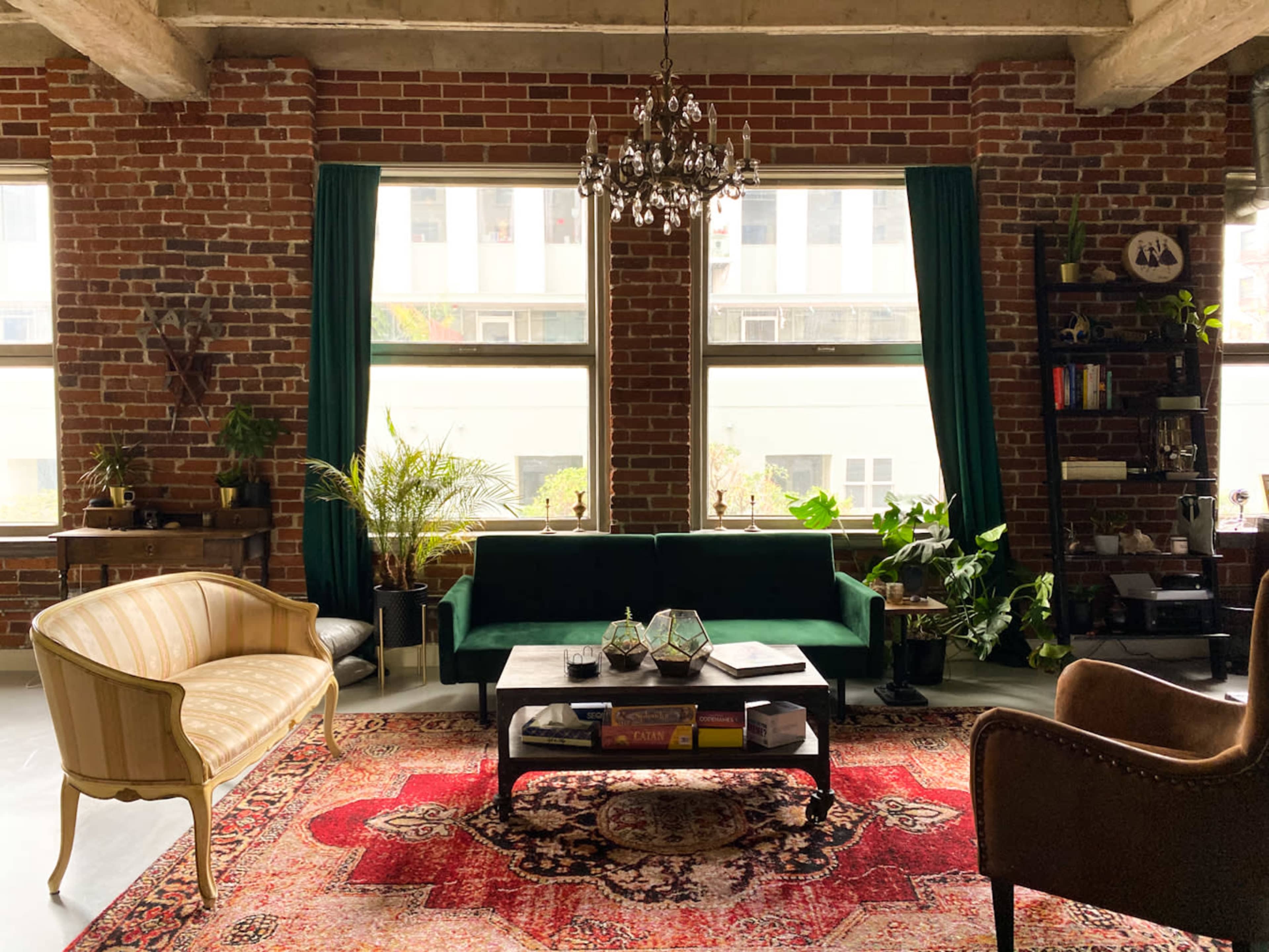 A cozy living room features a green sofa, a patterned rug, and large windows surrounded by brick walls and indoor plants.