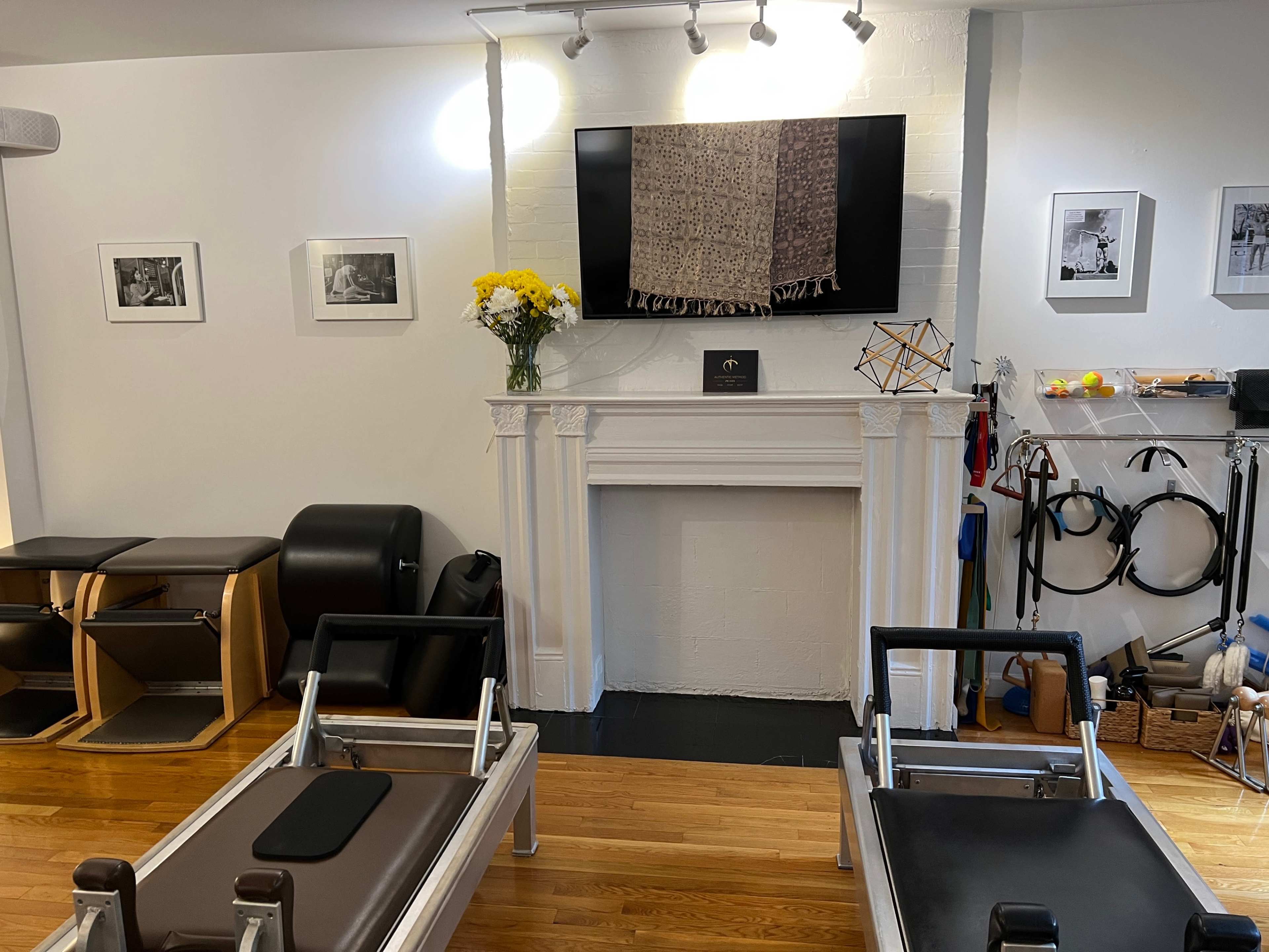 The image shows a fitness studio with two reformer machines, a wall-mounted television, and decorated with framed pictures and a flower arrangement on a mantelpiece.