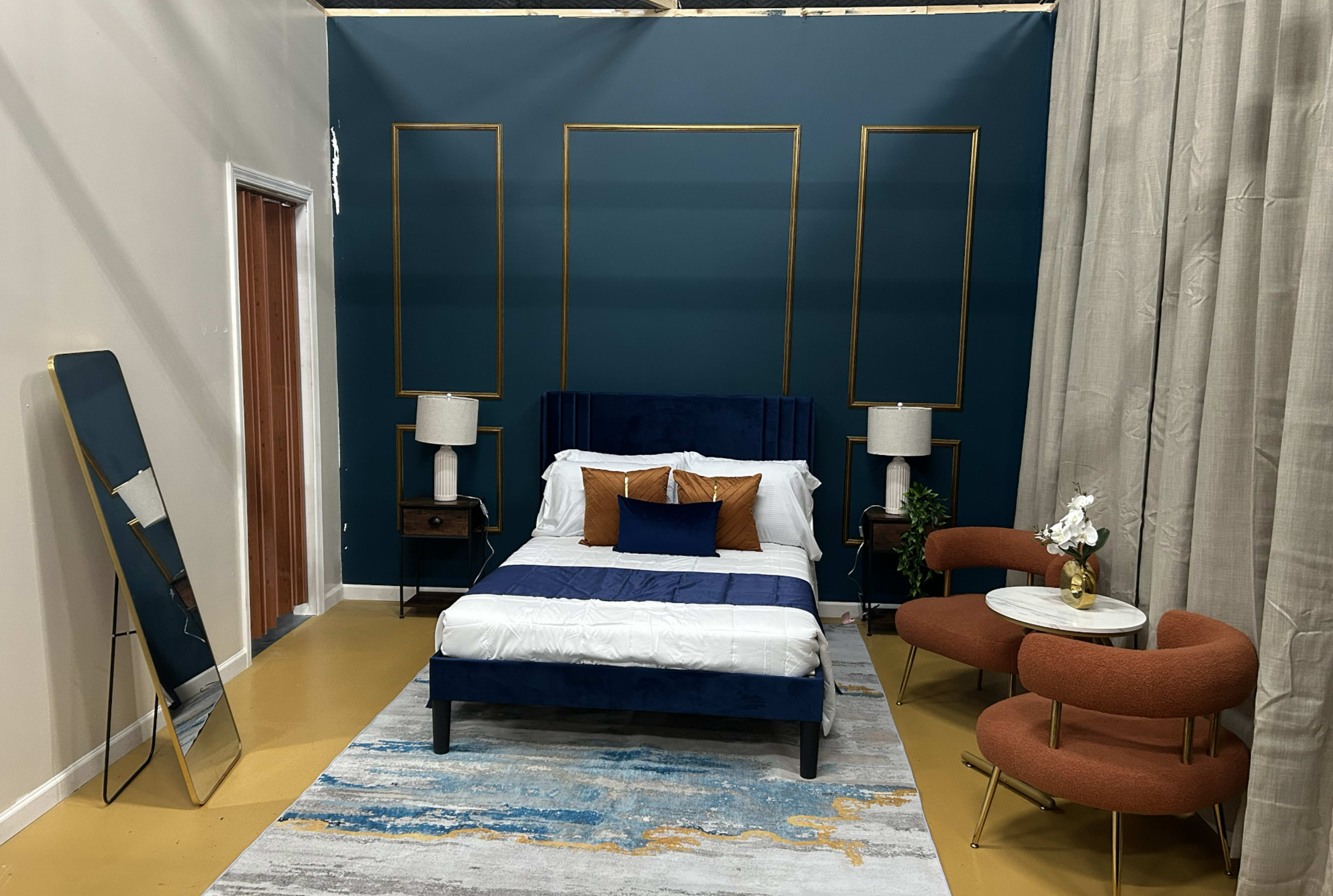 The image shows a styled bedroom featuring a bed with blue upholstery, two bedside lamps on wooden tables, a rug, a mirror, and two orange armchairs against a teal wall.