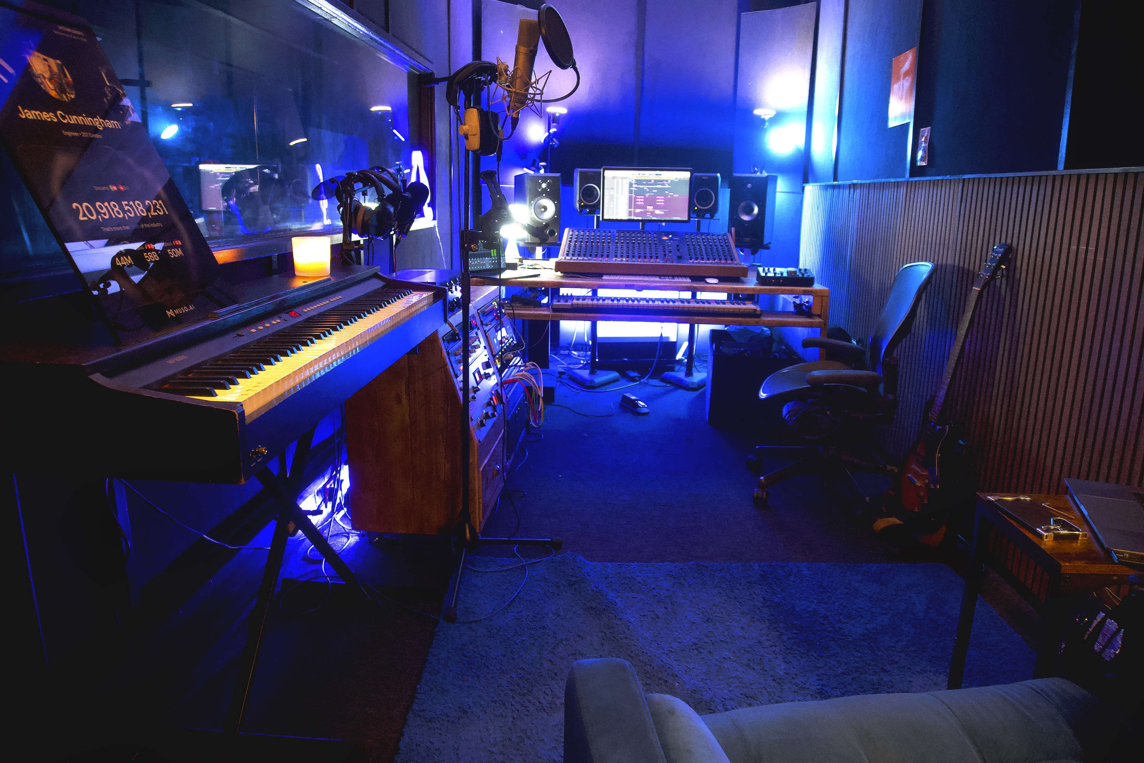 The image shows a music studio featuring a keyboard, audio equipment, a computer setup, and ambient lighting.