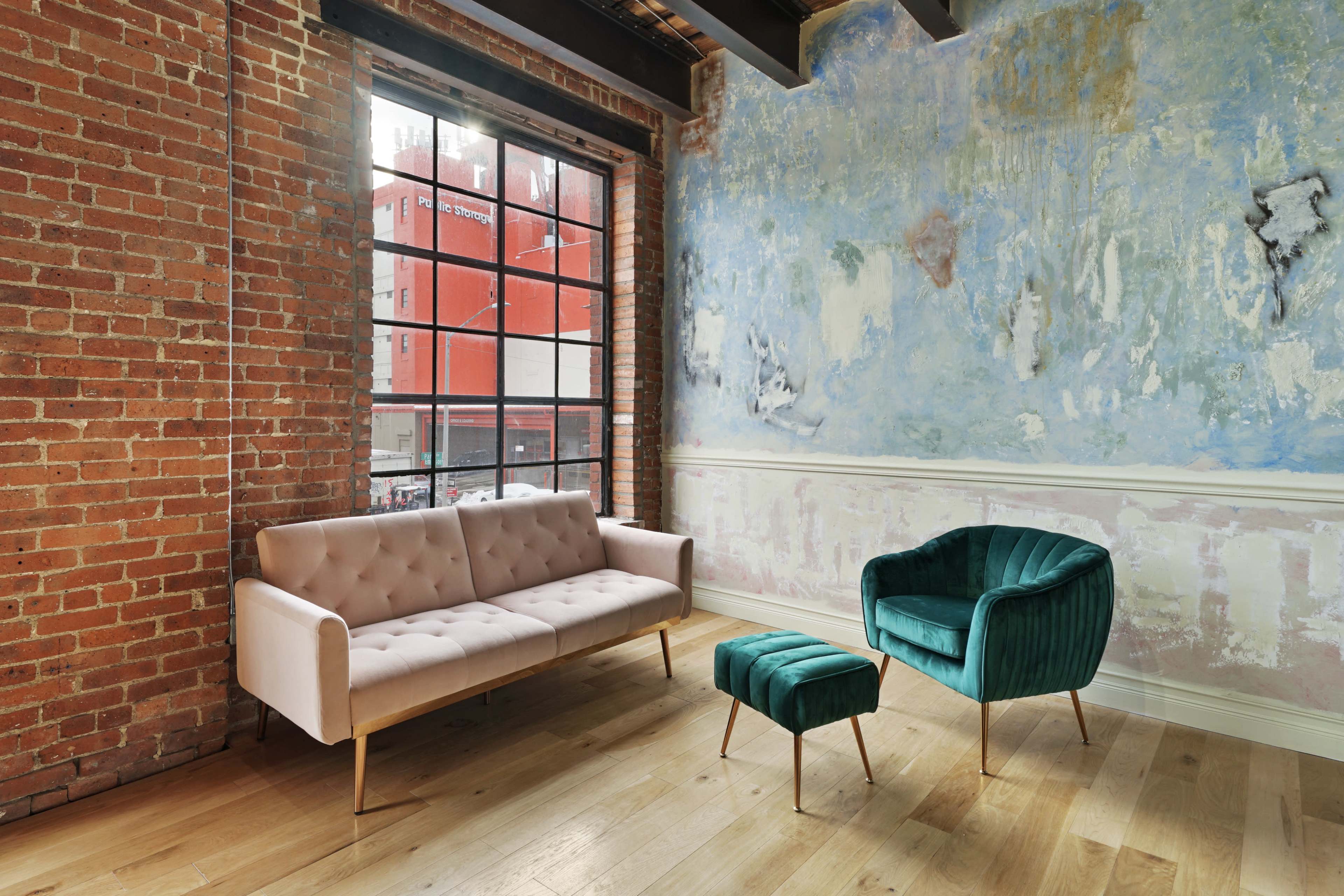 A living space with a pink tufted sofa, a green velvet armchair, and a matching ottoman, set against a backdrop of exposed brick walls and a painted blue wall with large windows.