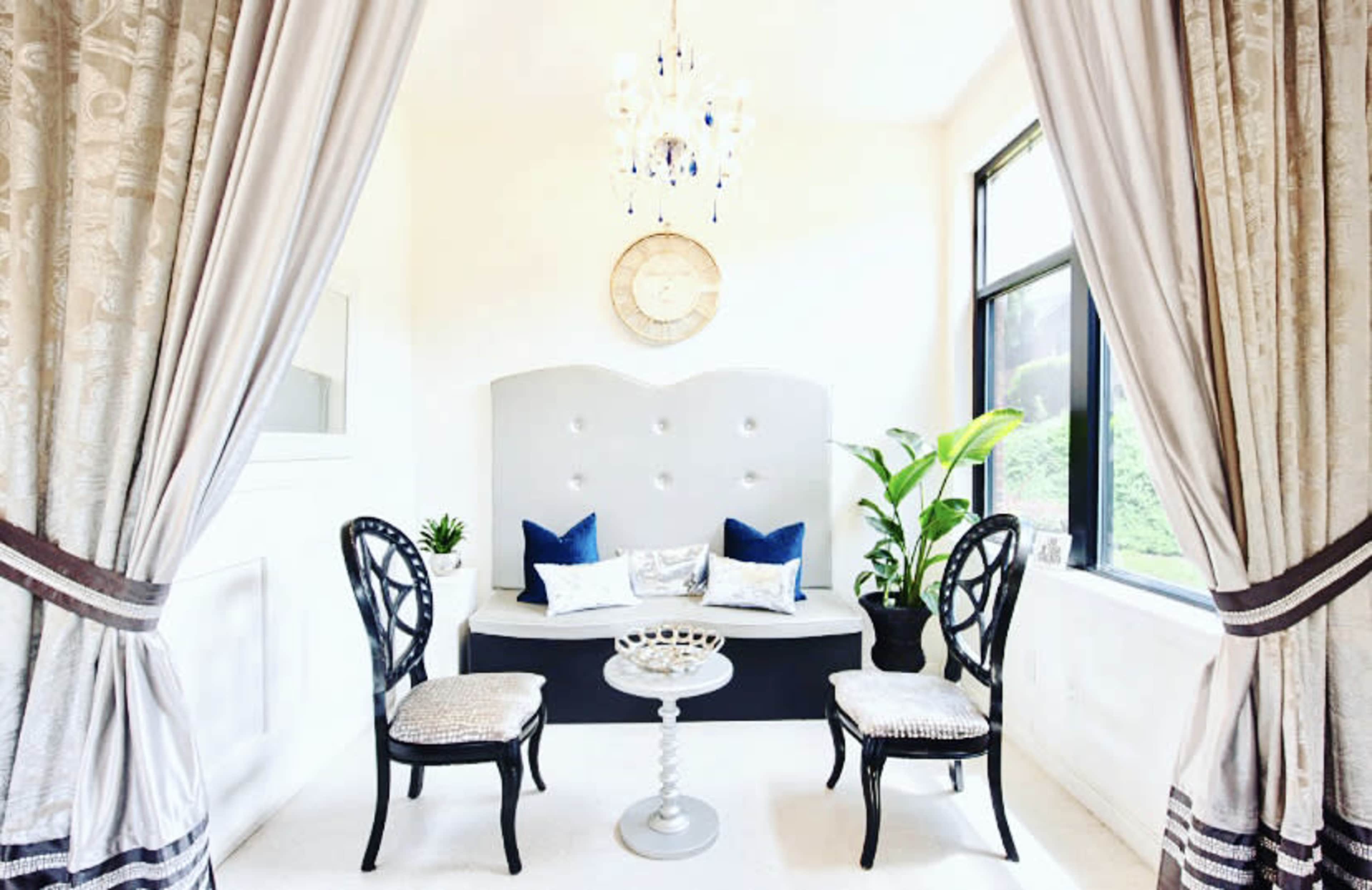 The image shows a small sitting area with a gray upholstered bench, two black chairs, a round table, and a plant, all framed by elegant curtains.