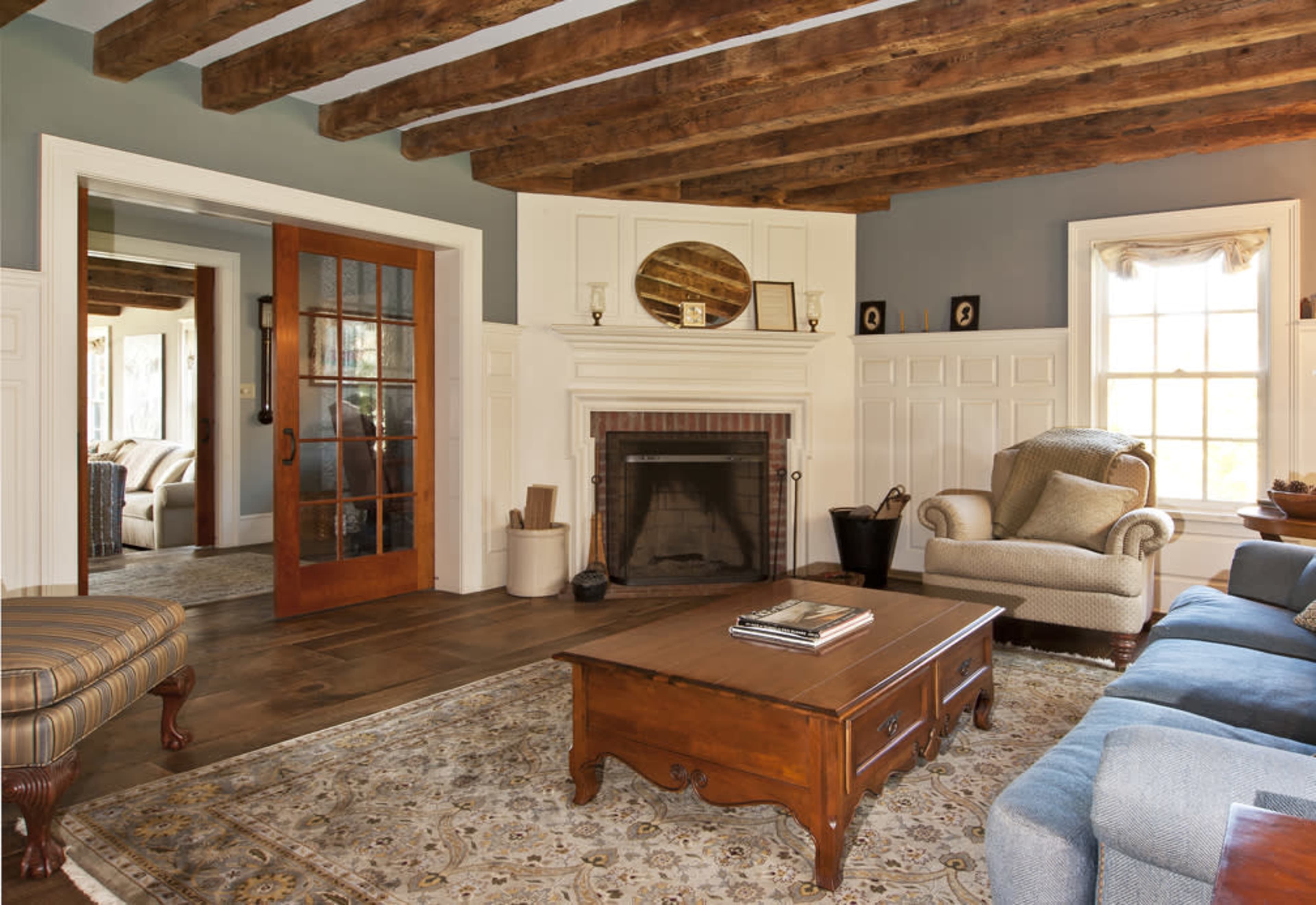 The living room features exposed wooden beams, a fireplace with a brick surround, a wooden coffee table, and a comfortable seating arrangement.