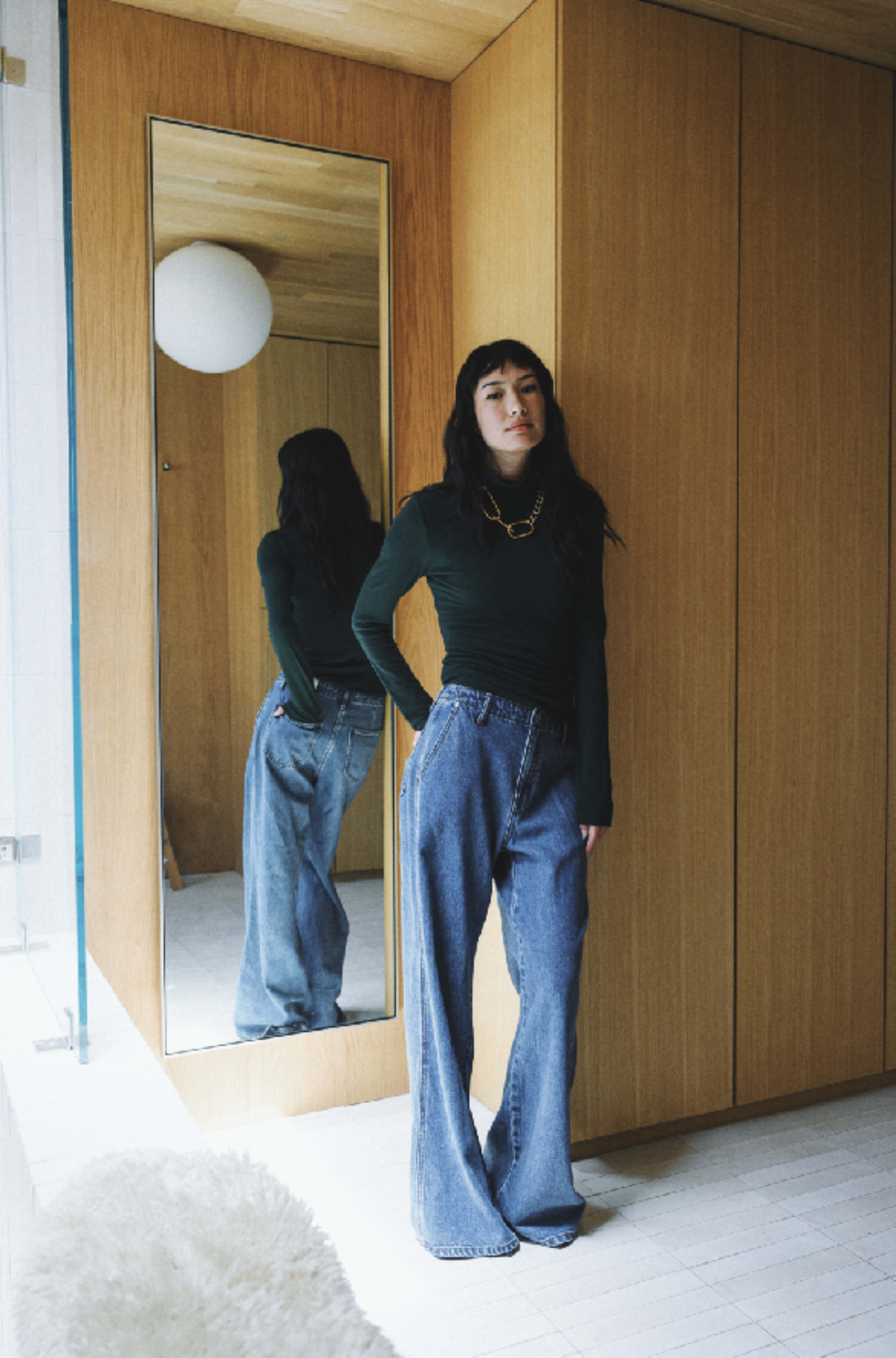 A person stands near a large mirror in a wooden-paneled room, wearing a green long-sleeve top and wide-legged jeans.