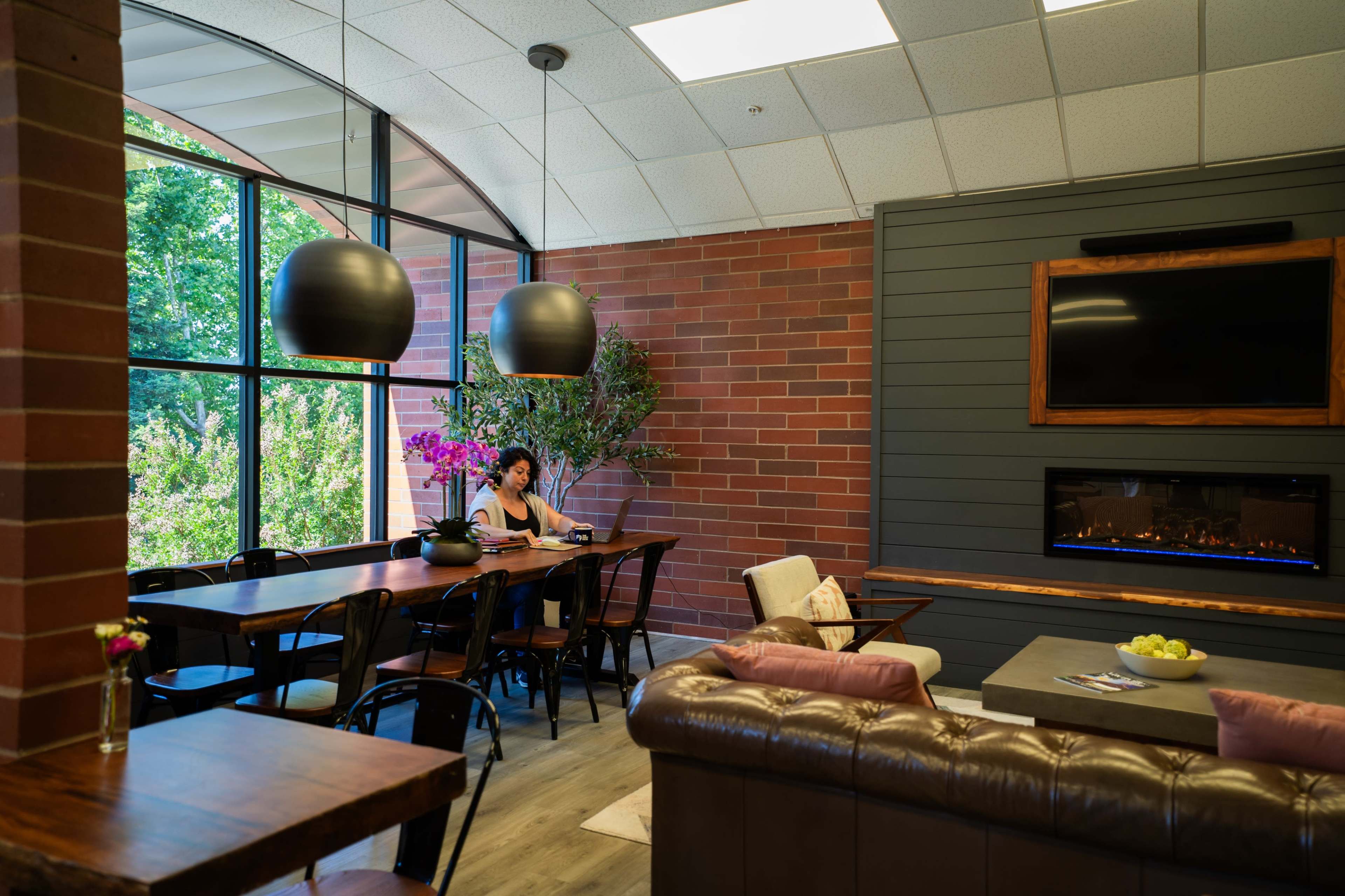 A woman sits at a long wooden table in a modern room featuring large windows, a gas fireplace, and comfortable seating.