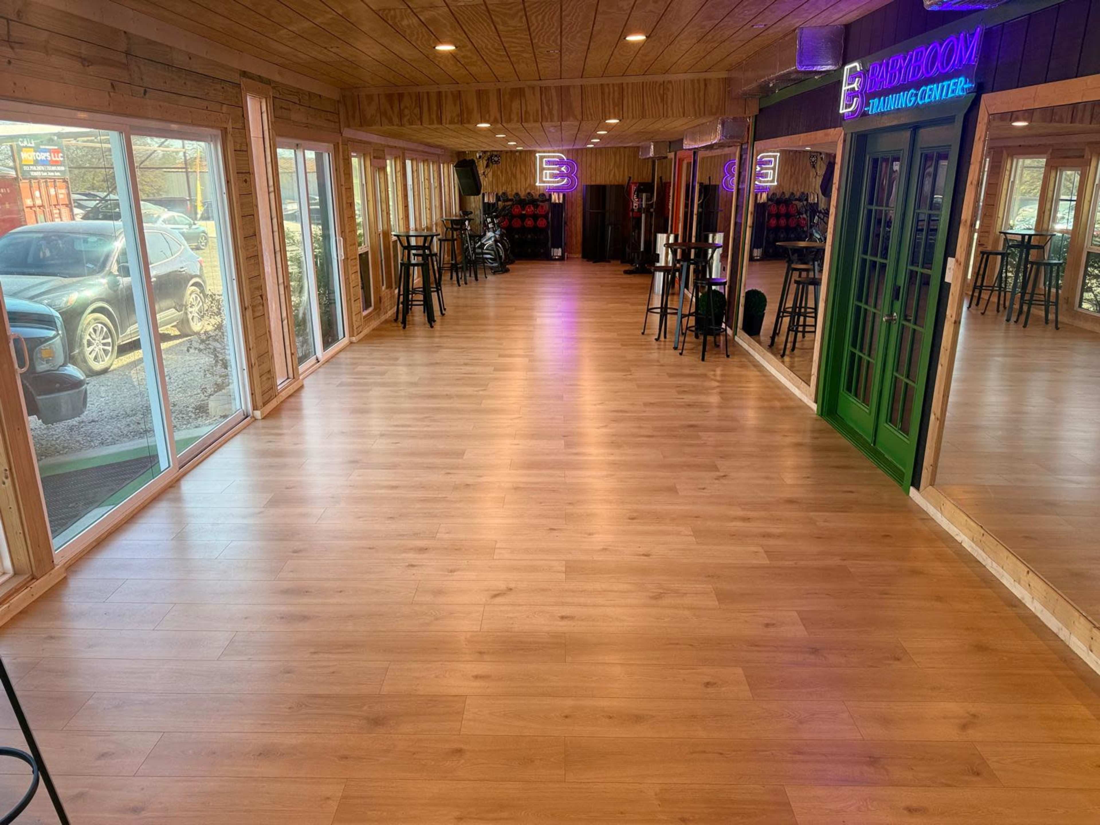 The image shows a spacious wooden interior with large windows, tables, and chairs, featuring a neon sign that reads "E" and a green door.