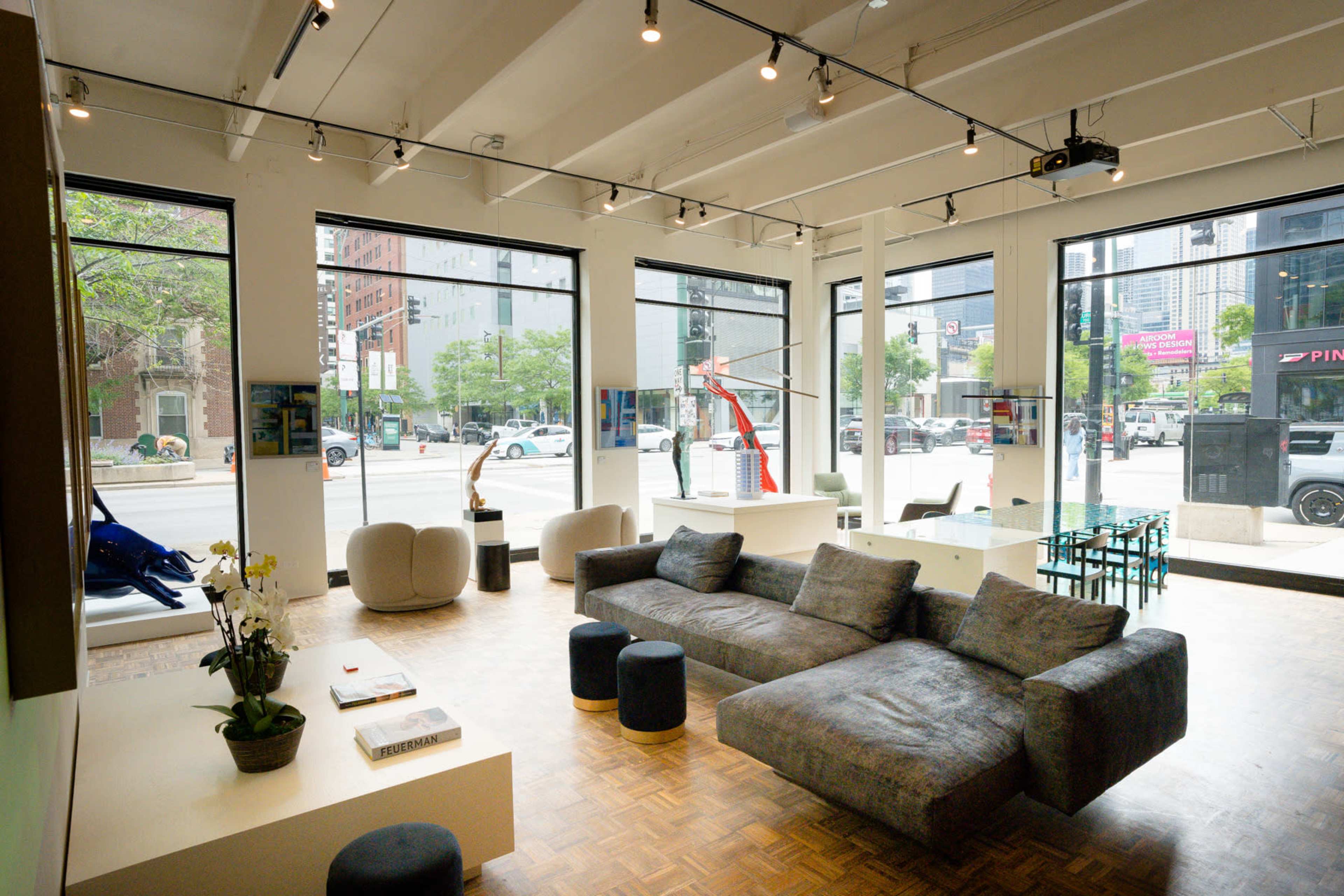 The image shows a modern, spacious open area with large windows, featuring a minimalist seating arrangement, a coffee table, and a dining table near the street.