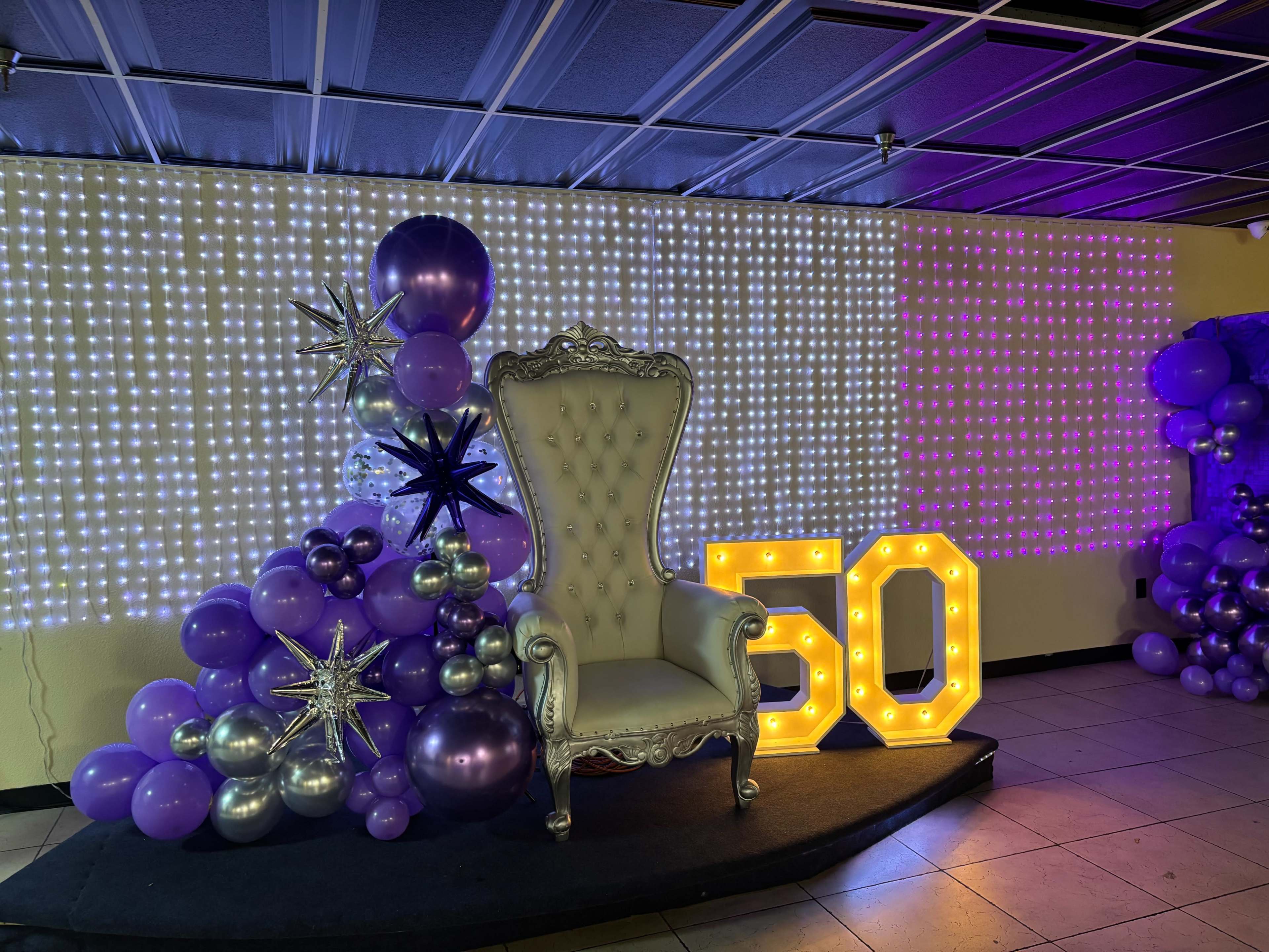A lavish white throne is centered in front of a twinkling backdrop of lights, surrounded by purple balloons and decorative elements, with a large illuminated "50" sign beside it.