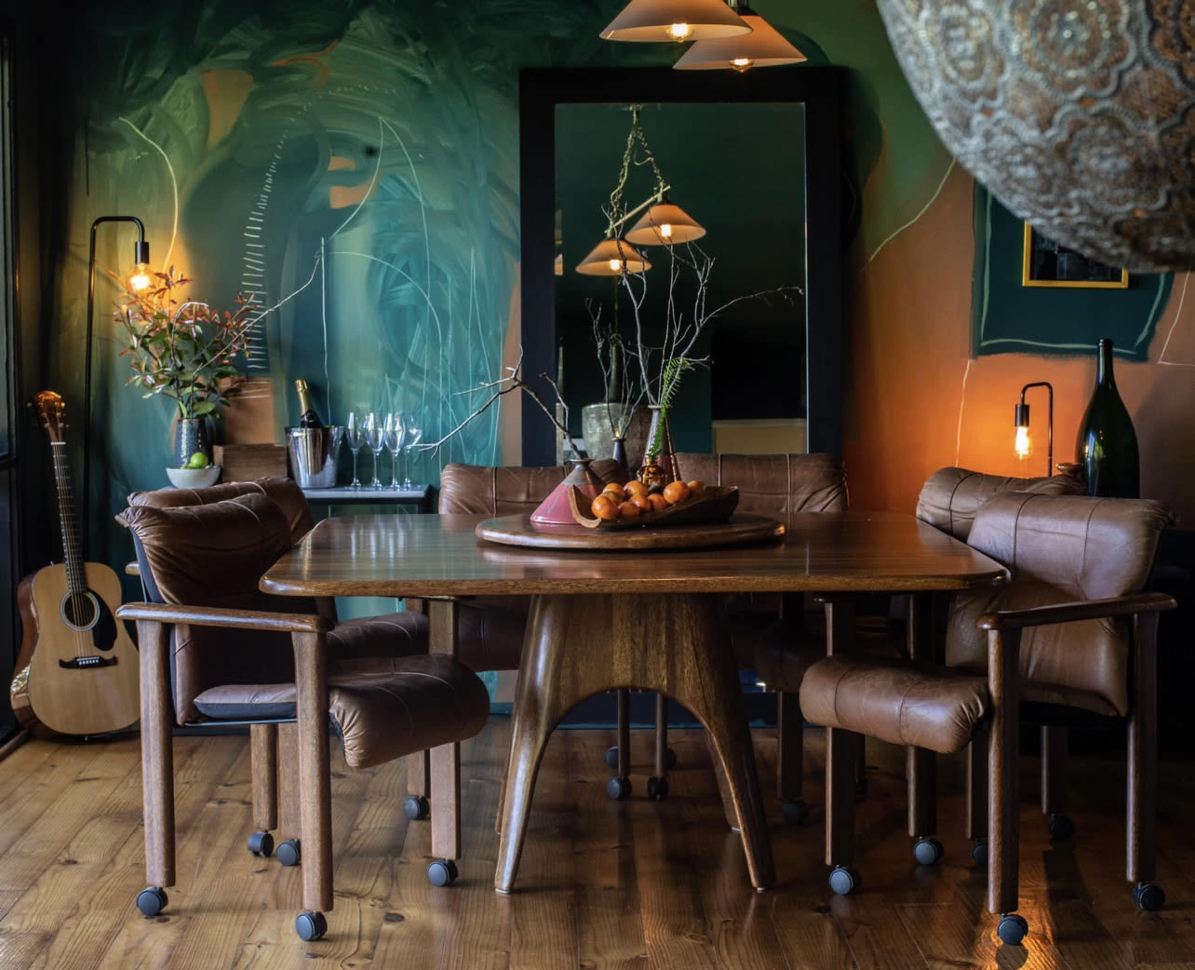 A mid-century modern dining room features a wooden table surrounded by leather chairs, with a large mirror and decorative elements against a colorful mural backdrop.