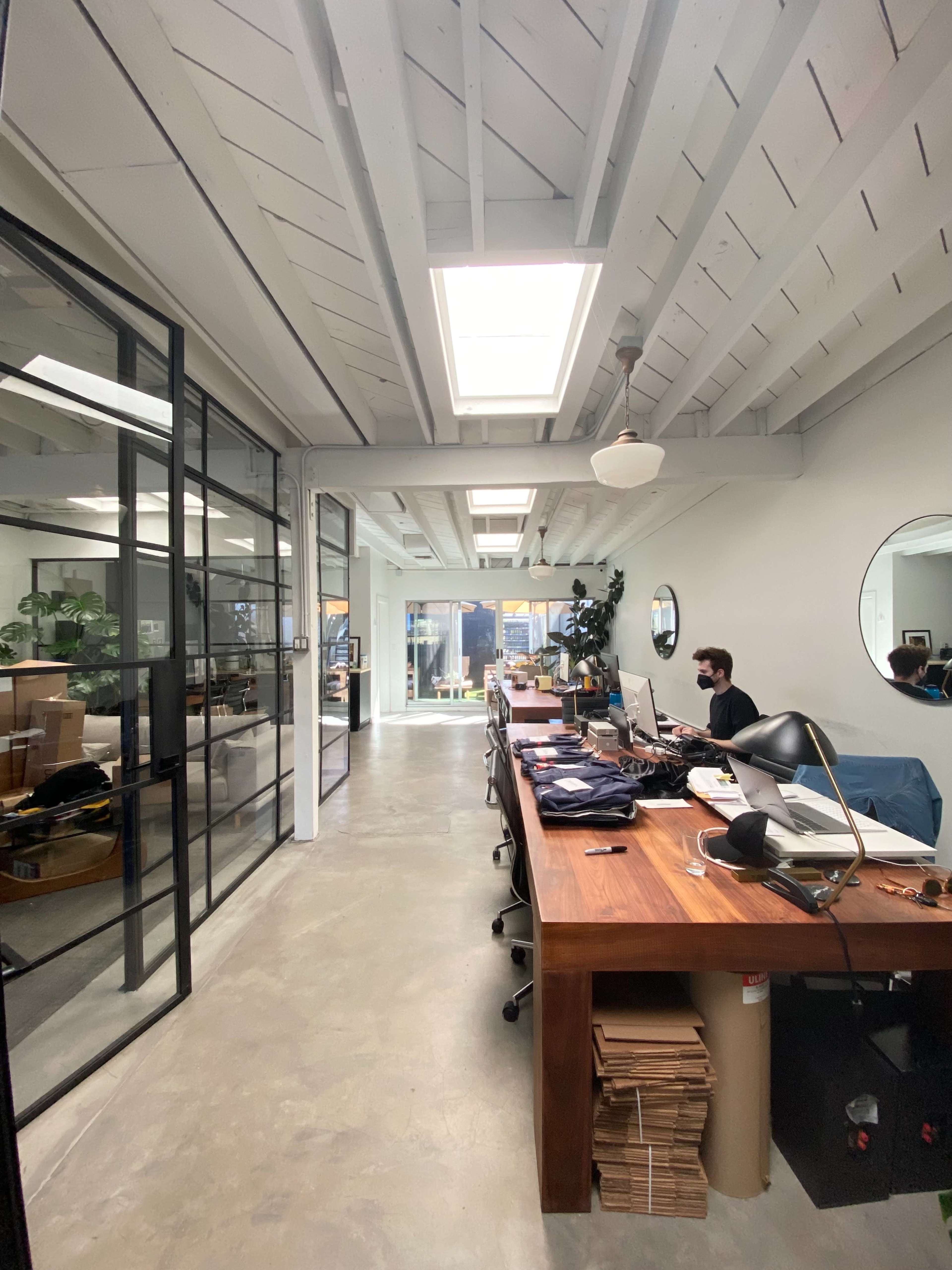 A spacious office area features a long wooden table with scattered items, large windows allowing natural light, and a visible workspace with a man seated at a desk.