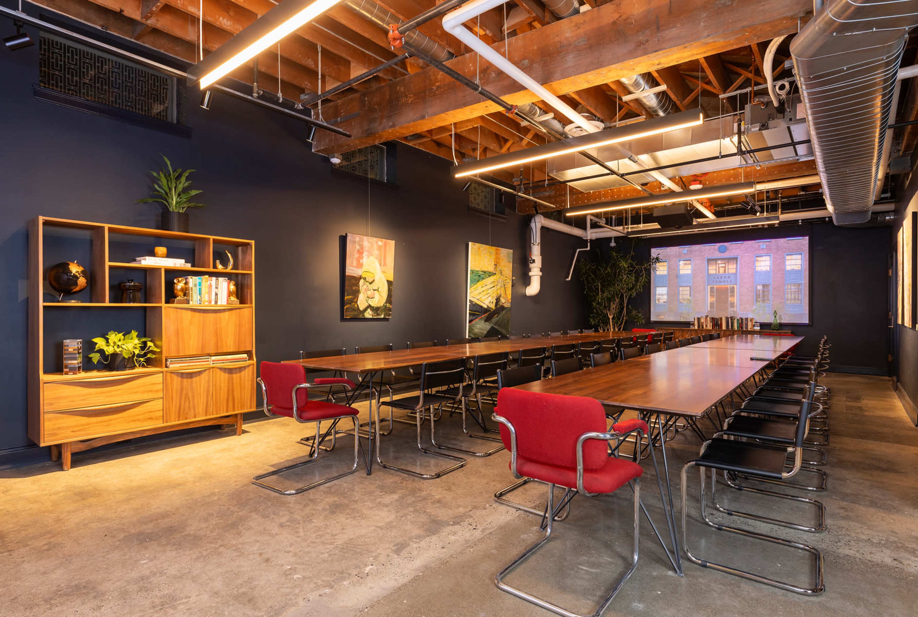 A conference room features long wooden tables with red chairs, a wooden shelving unit, and large windows displaying an outdoor view.