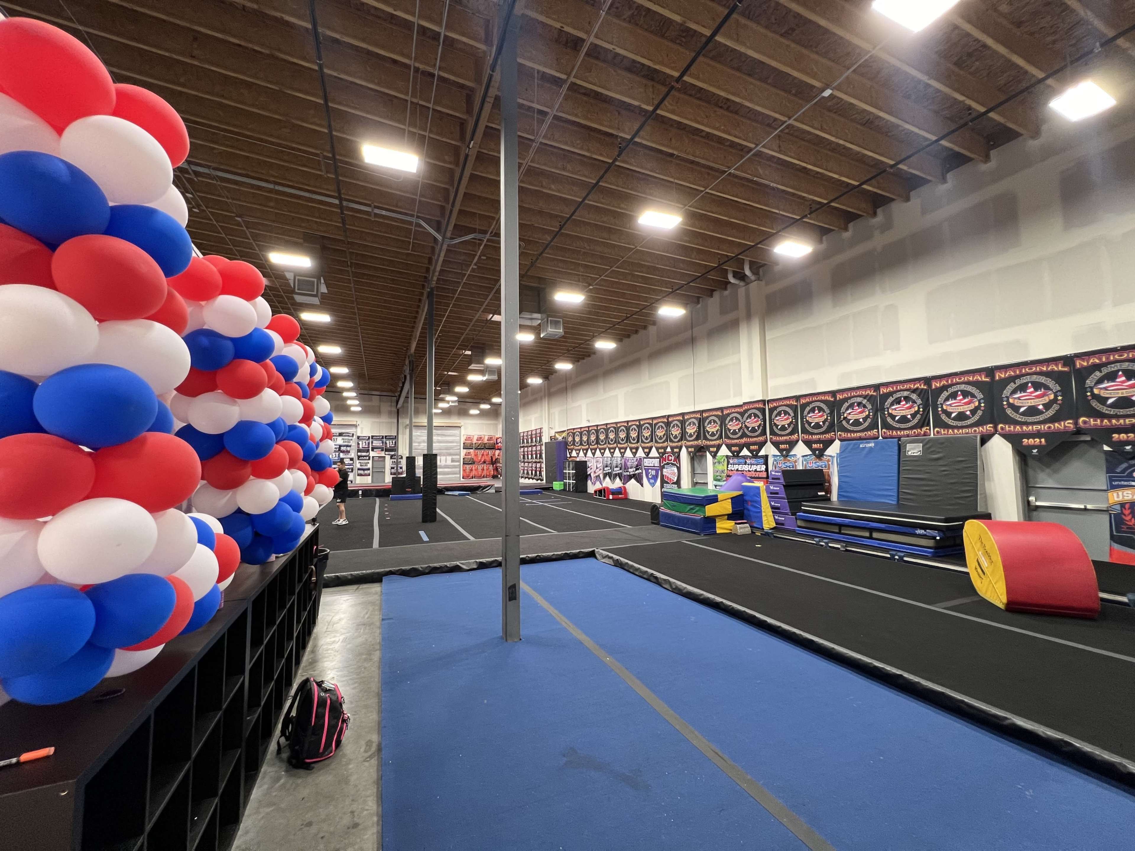 An indoor gymnastics facility featuring various exercise mats, equipment, and a decorative wall of red, white, and blue balloons.