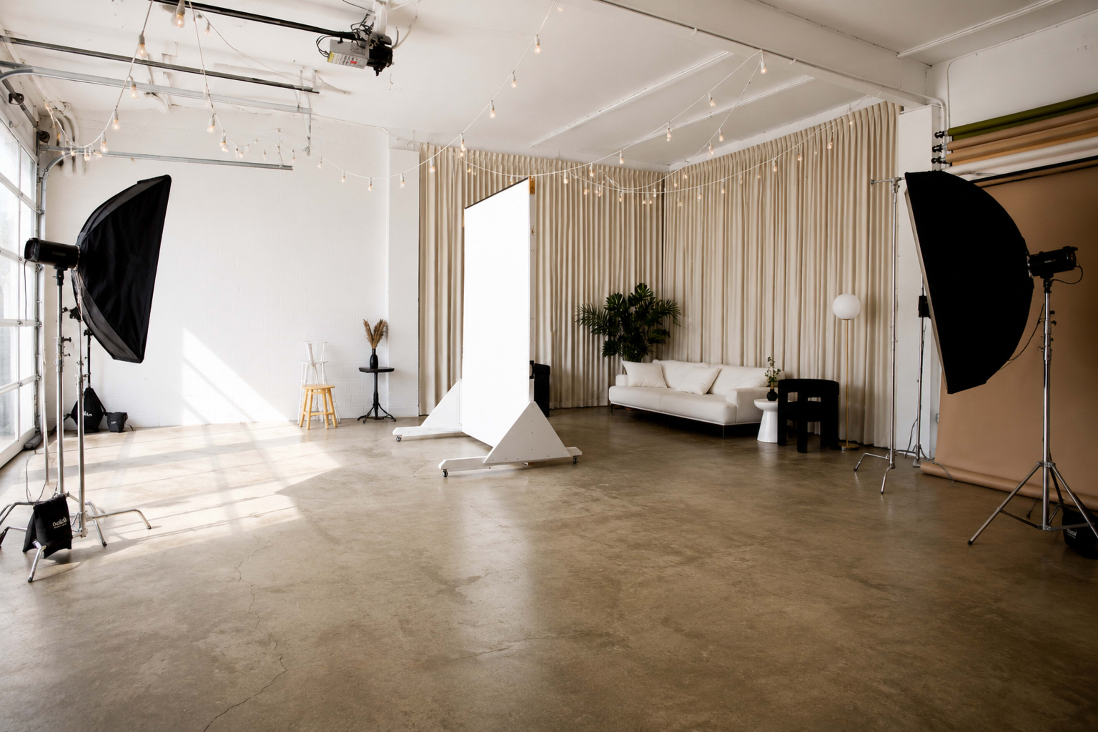 The image shows a photography studio with a large white backdrop, two lighting setups, a couch, and a decorative plant.