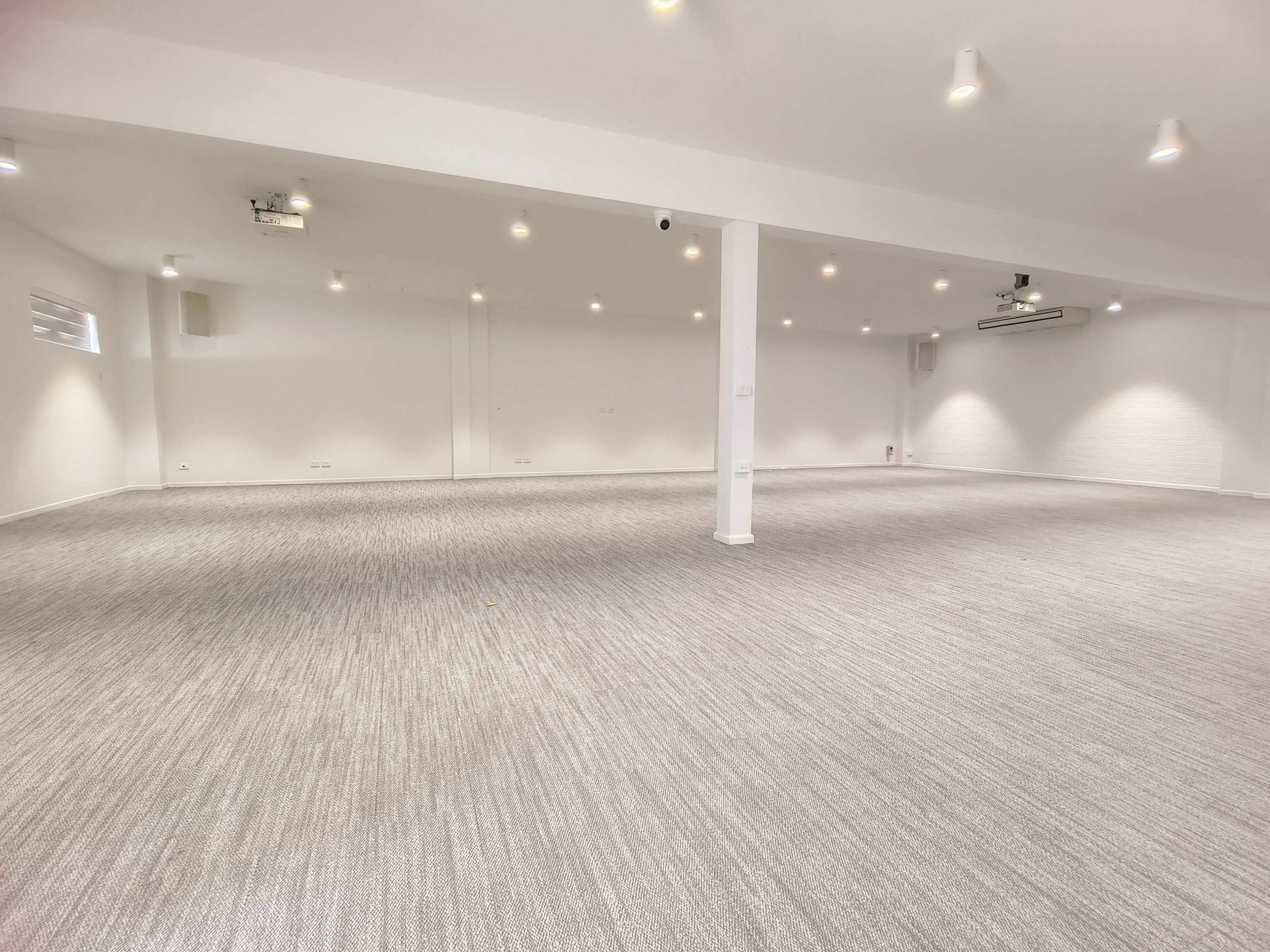 The image shows a large, empty interior space with white walls and a carpeted floor, illuminated by ceiling lights.