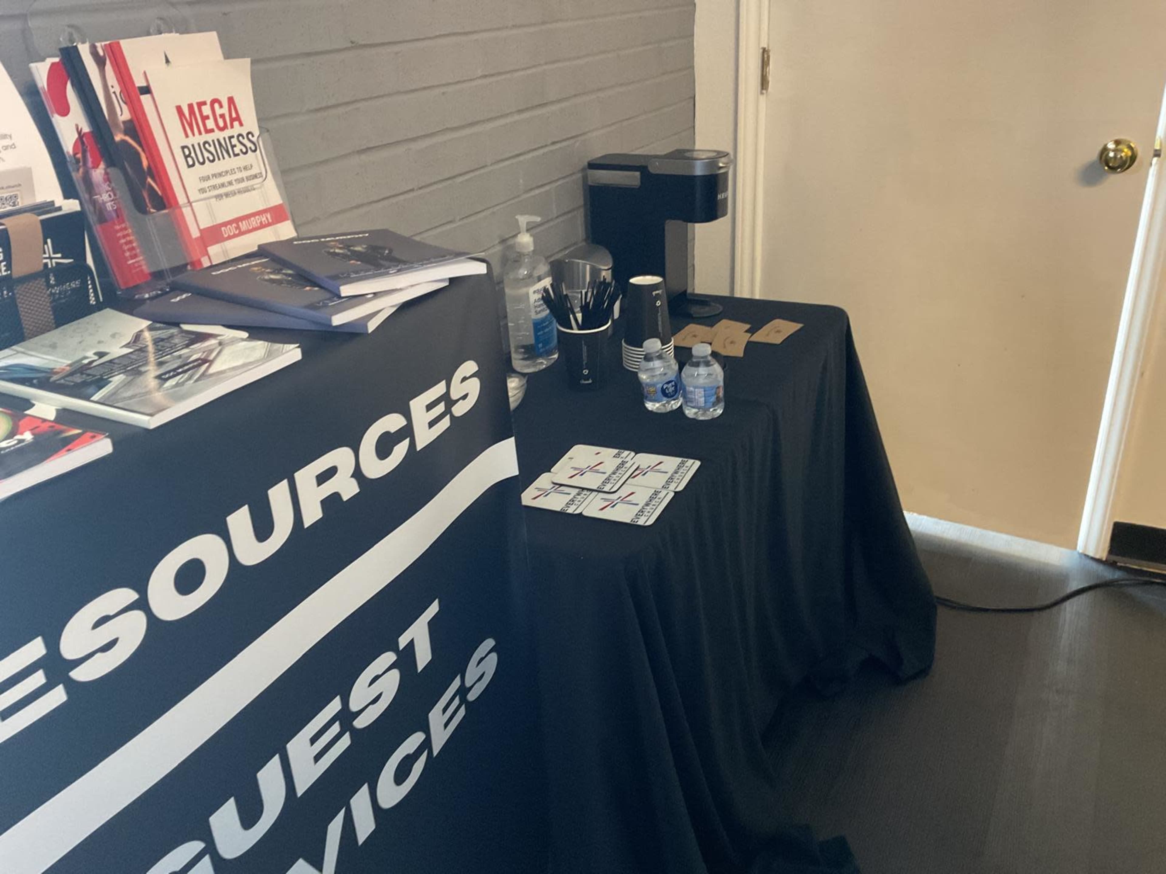 A black table with a tablecloth displays brochures and a coffee maker next to bottled water and coasters.
