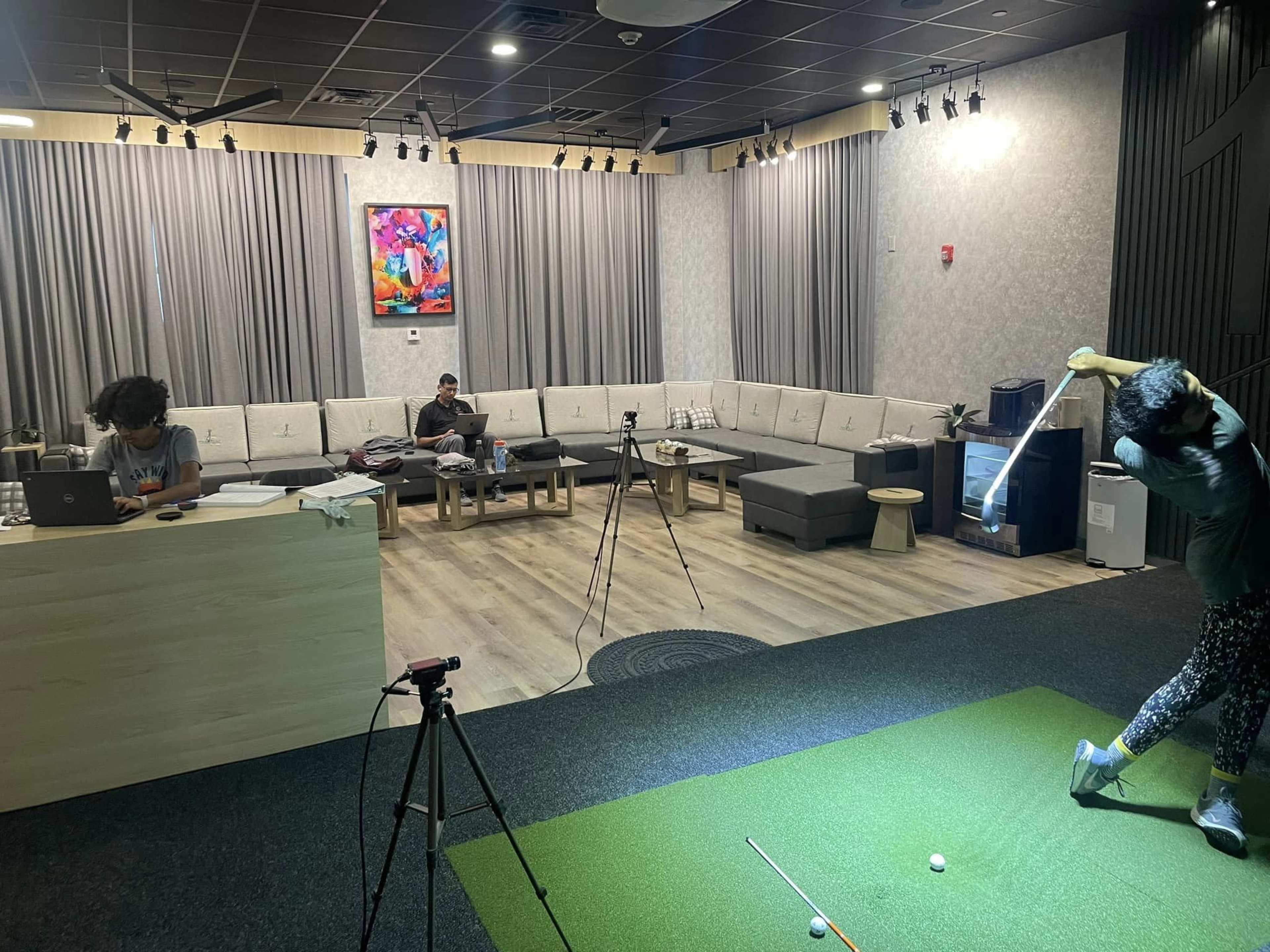 A man is practicing his golf swing on a green mat while another person sits on a couch with a laptop in a modern room with gray curtains.