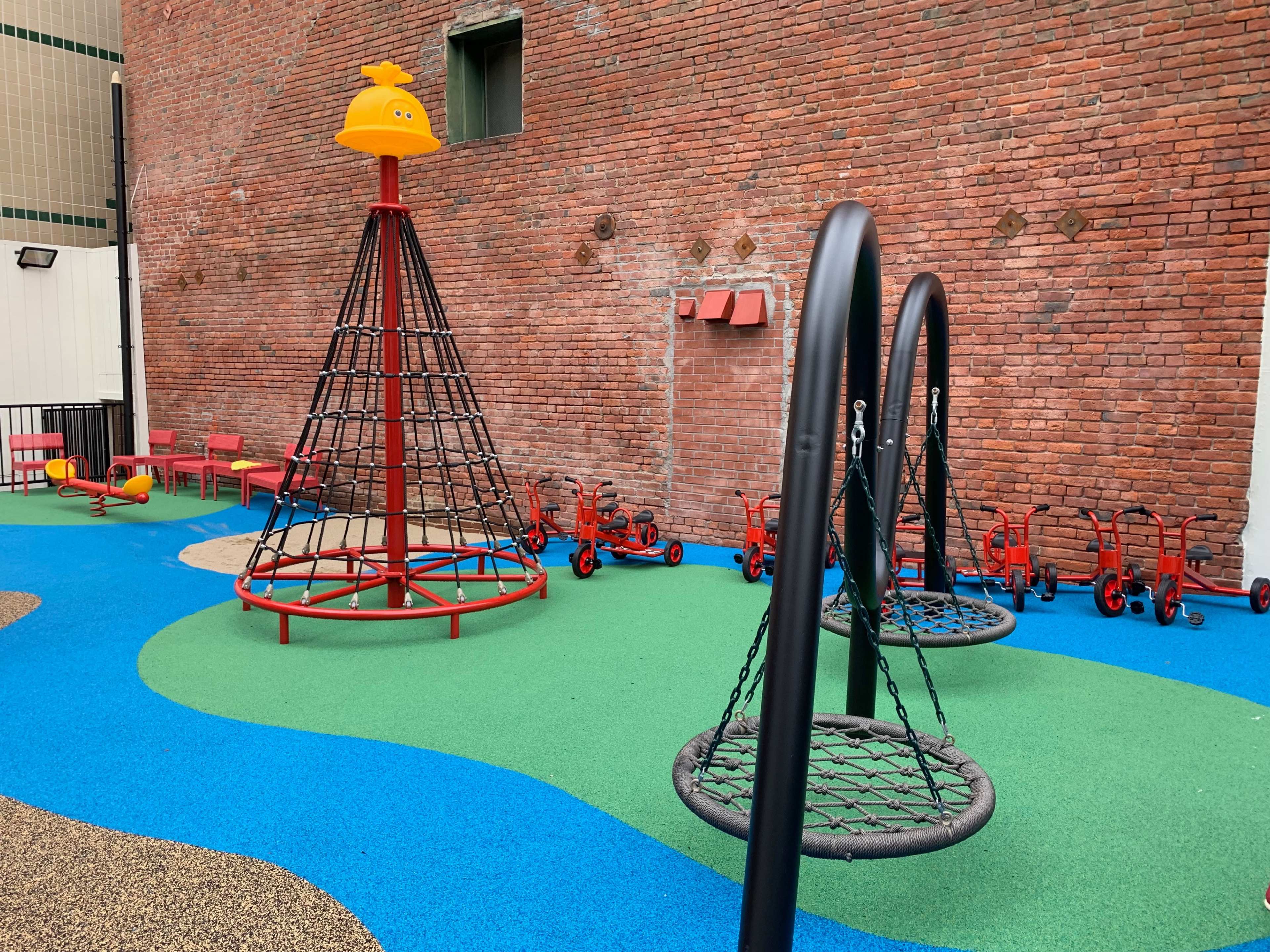 A playground with a climbing structure, swings, and red ride-on toys situated on a colorful rubber surface, surrounded by brick walls.