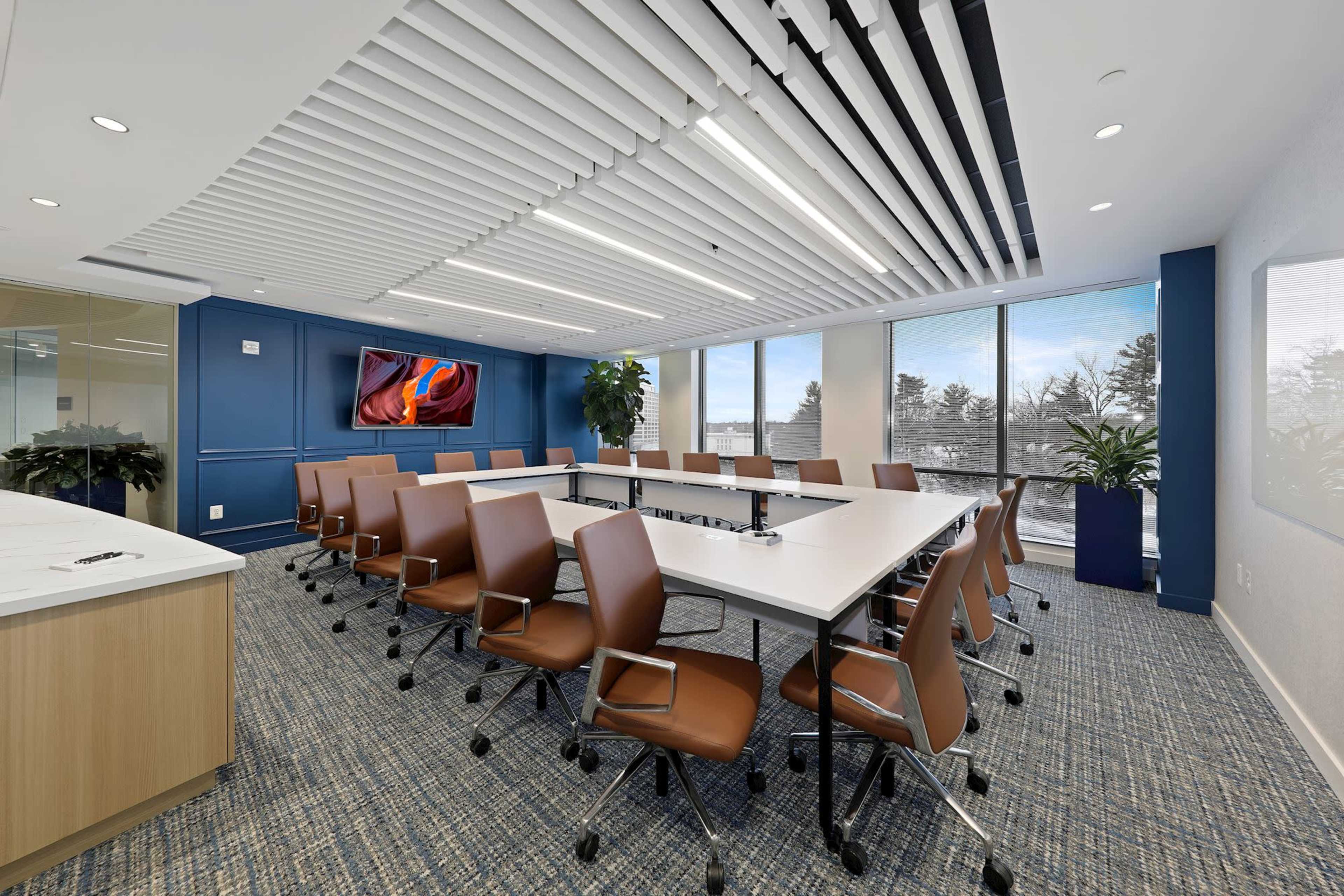 A modern conference room with a long white table surrounded by brown swivel chairs, large windows, and a wall-mounted screen.