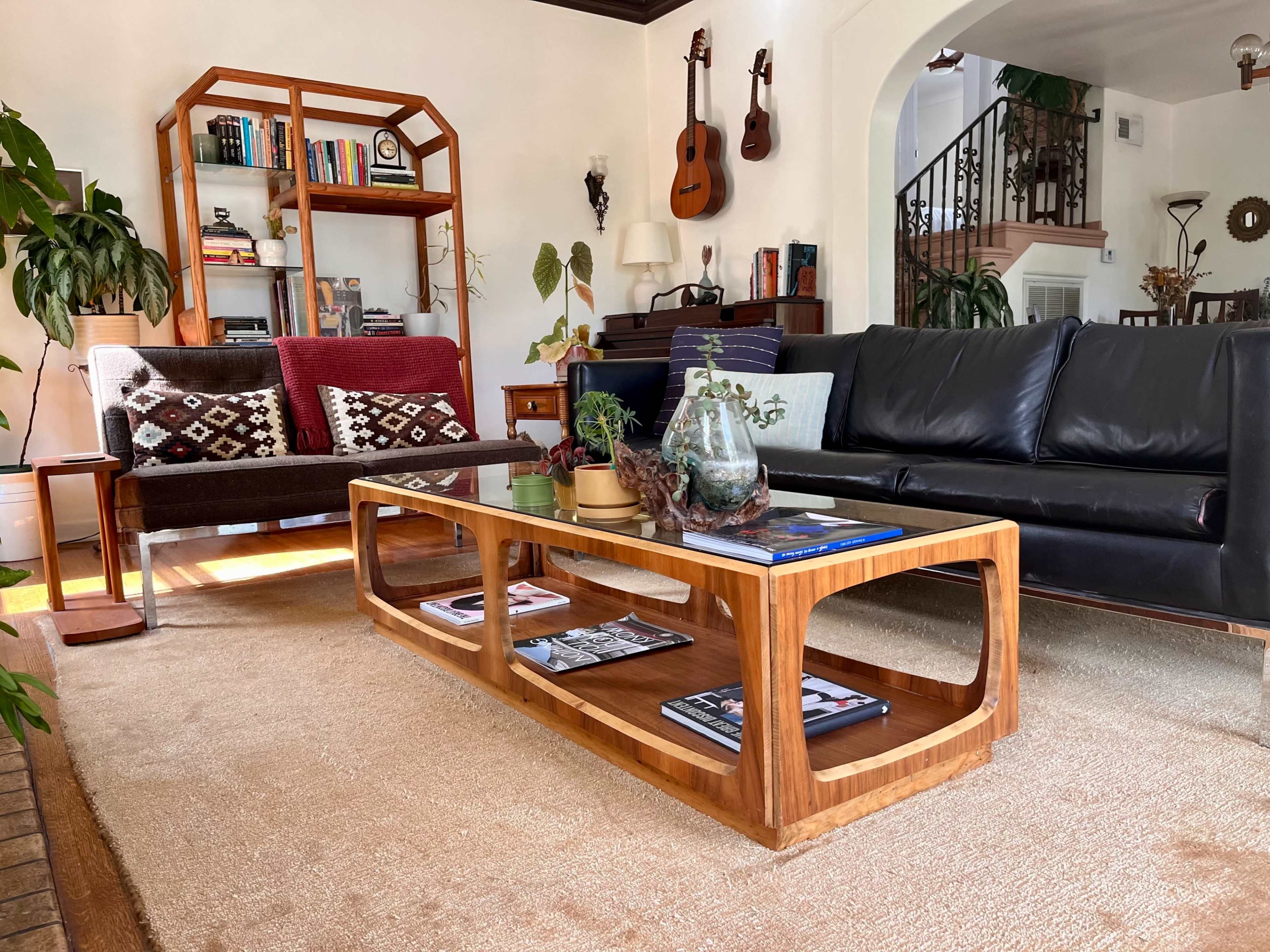 The image shows a cozy living room with a mix of modern and vintage furniture, including a wooden coffee table, two couches, a bookshelf filled with books, and decorative plants.
