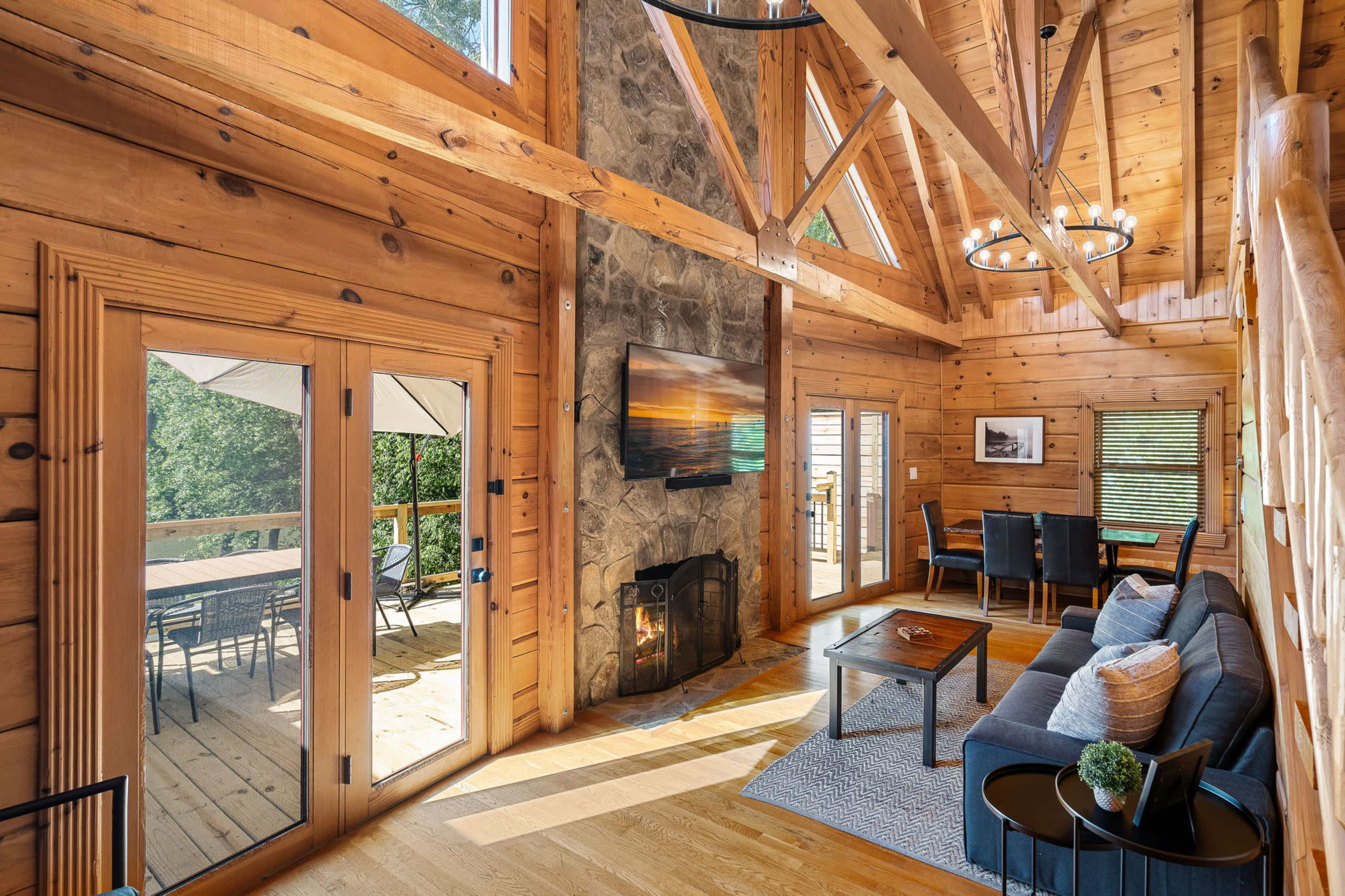 A cozy, wooden interior of a cabin featuring a stone fireplace, large windows, and a seating area with a view of an outdoor deck.