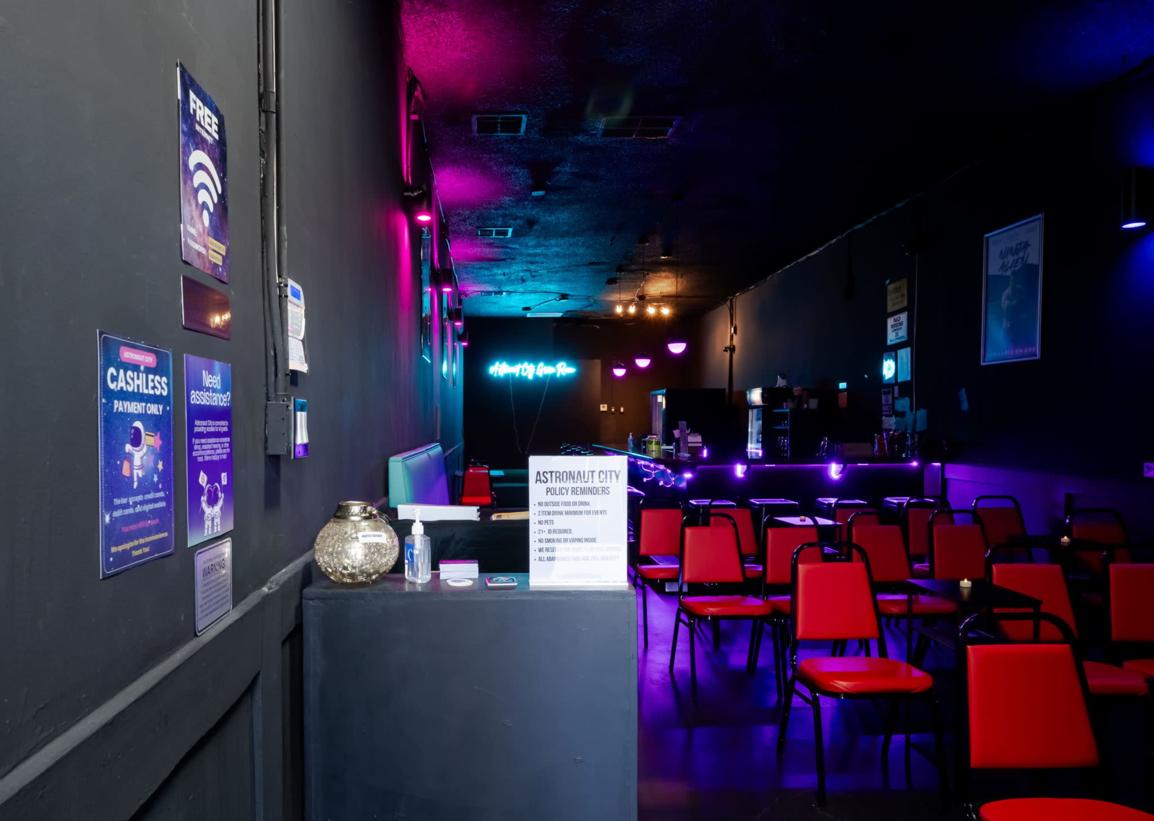 The interior of a dimly lit venue features a bar area with a sign, rows of red chairs facing a stage, and colorful lights illuminating the space.