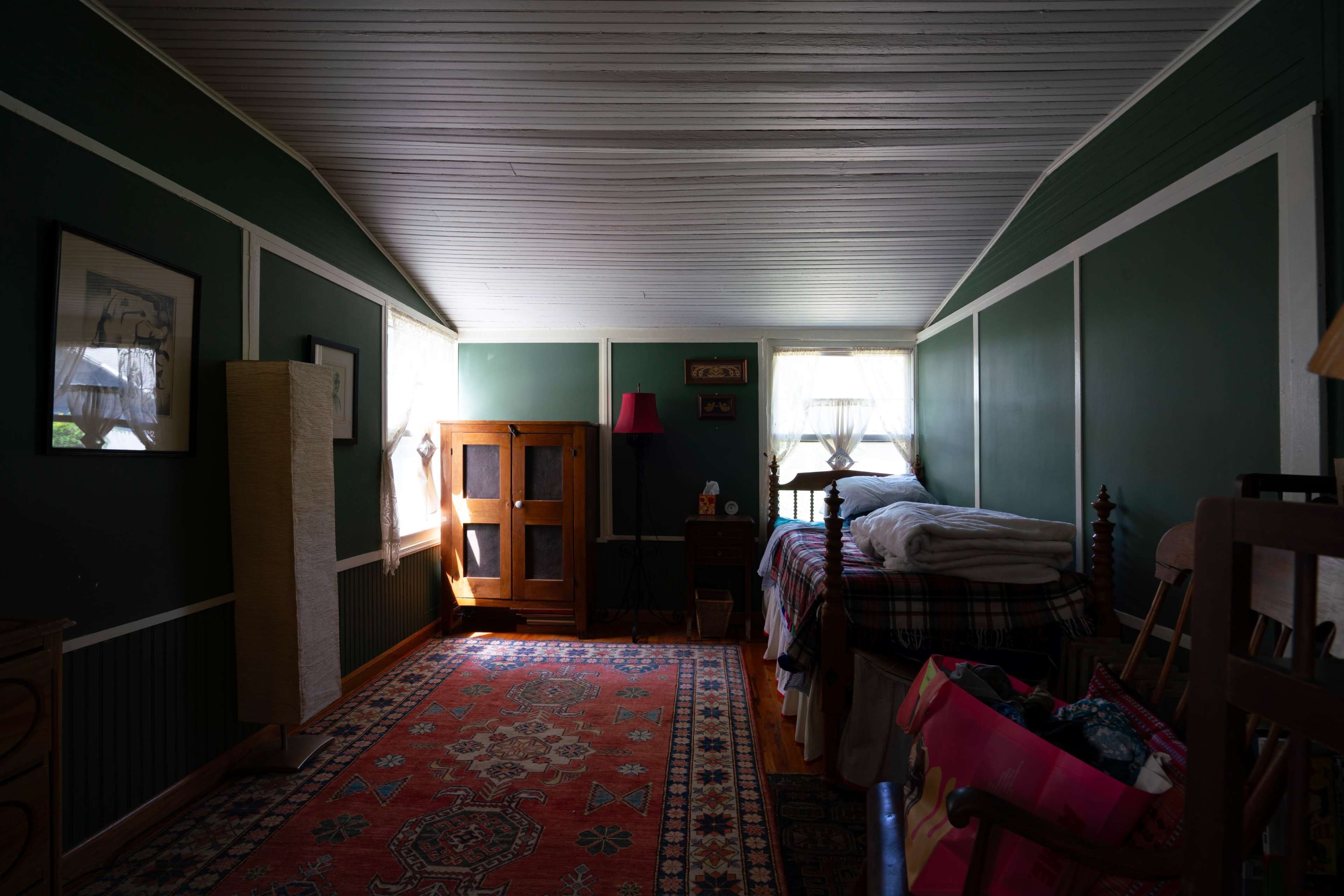 The image shows a room with green walls, a bed with a patterned blanket, a wooden dresser, and a patterned rug on the floor.