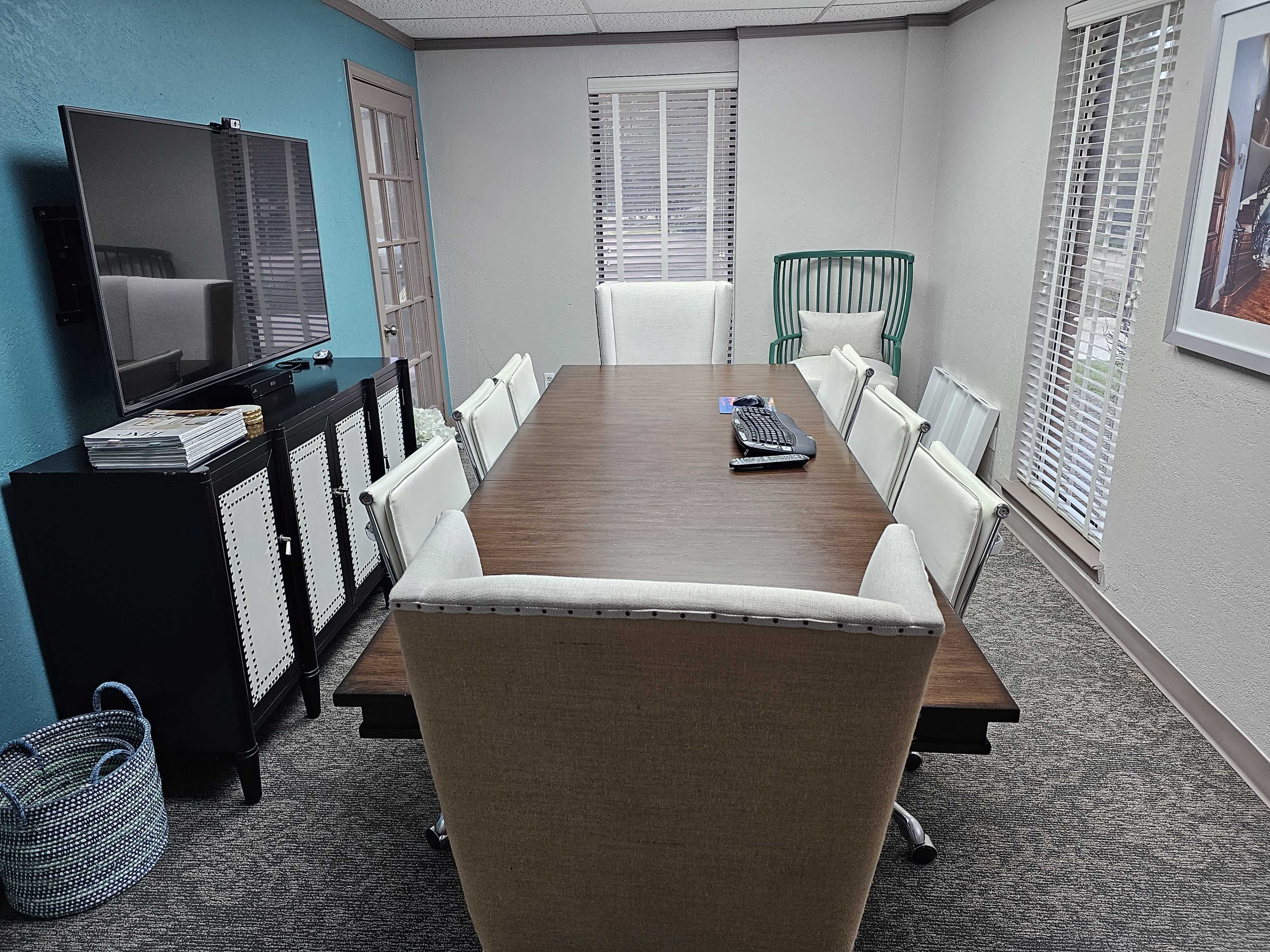 The image shows a modern conference room with a long wooden table surrounded by white chairs, a large television mounted on the wall, and a green accent chair in the corner.