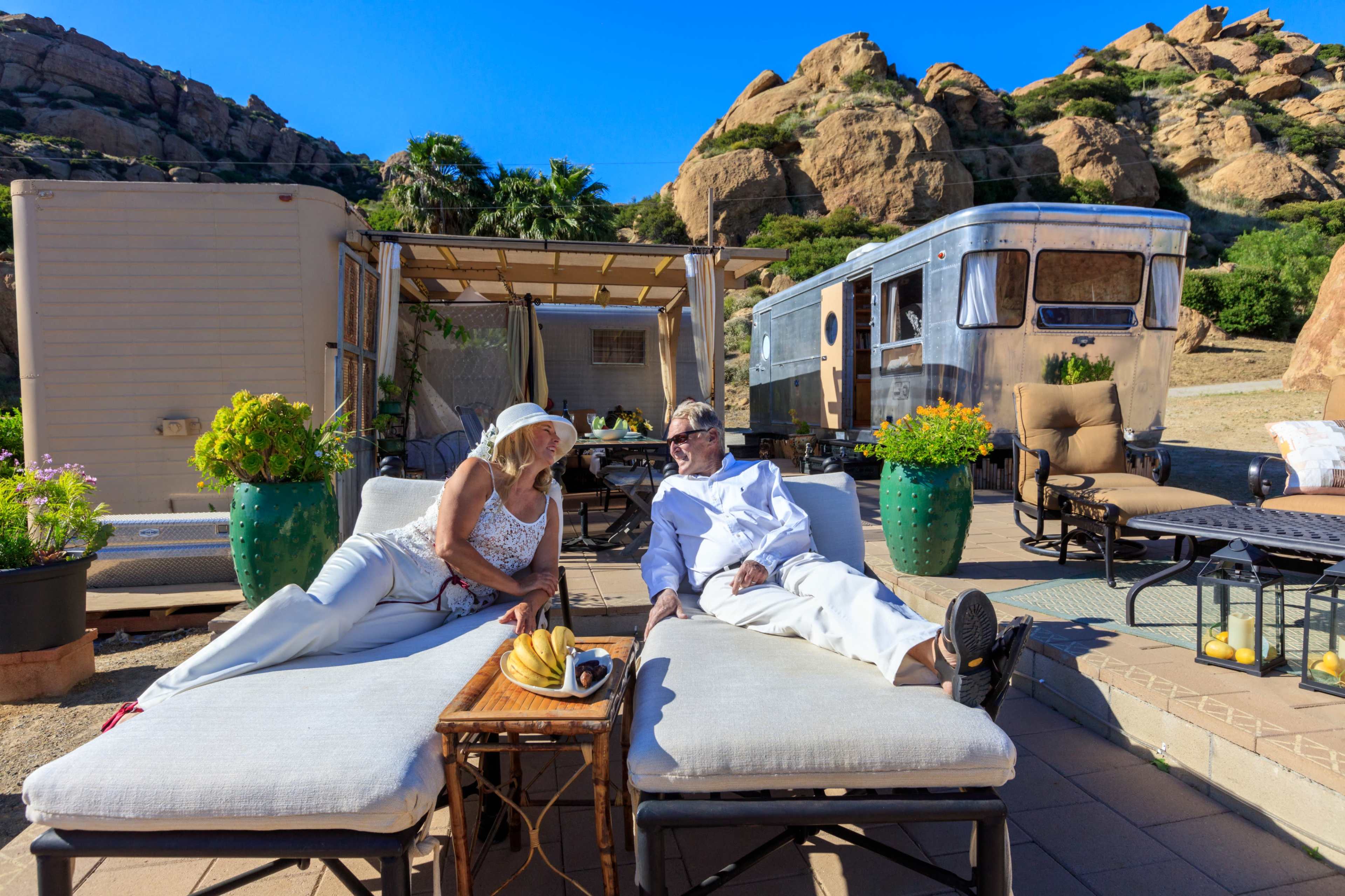 A couple relaxes on lounge chairs beside a vintage trailer in a desert landscape, surrounded by greenery and rock formations.