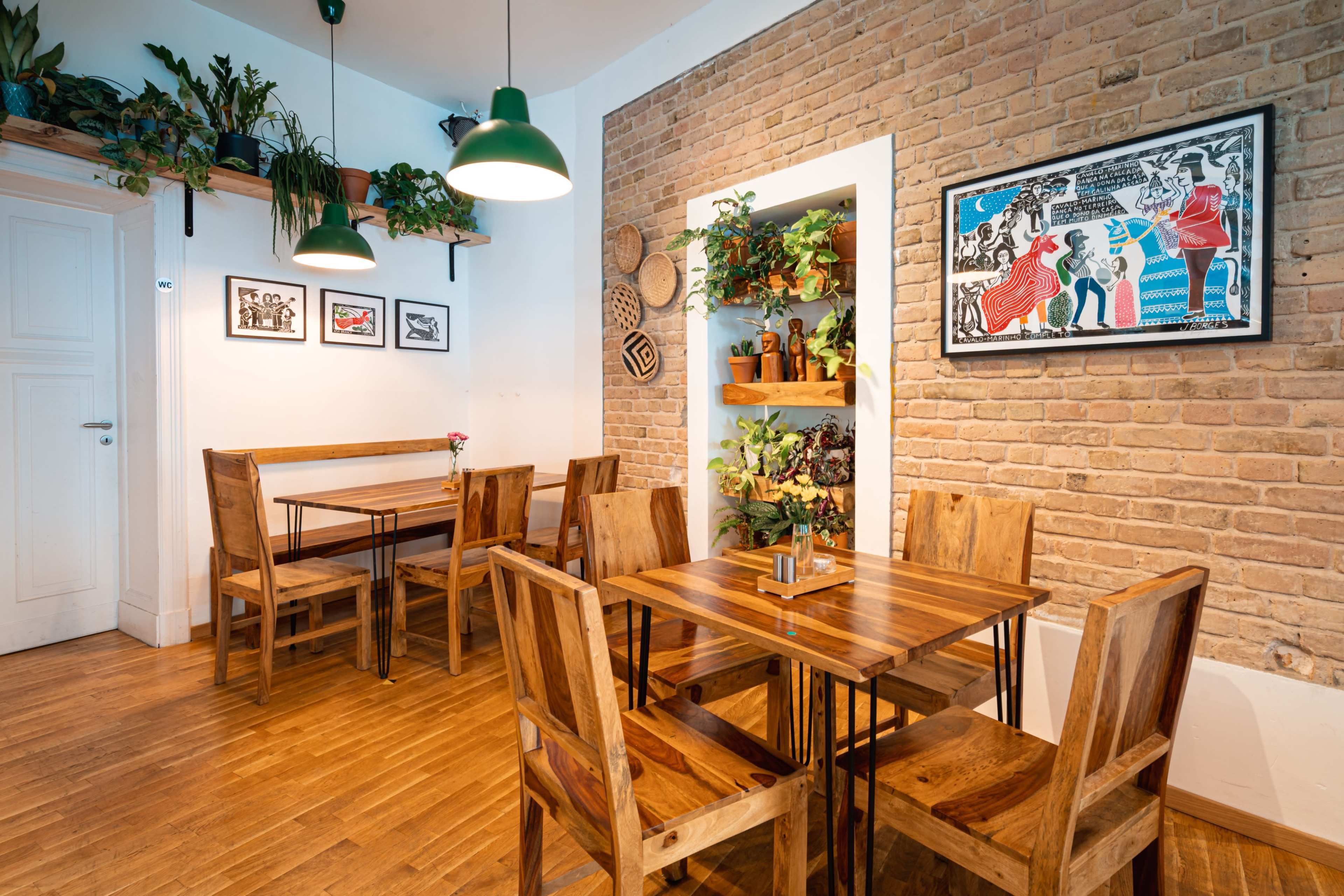 A cozy café interior featuring wooden tables and chairs, brick walls adorned with plants and artwork.