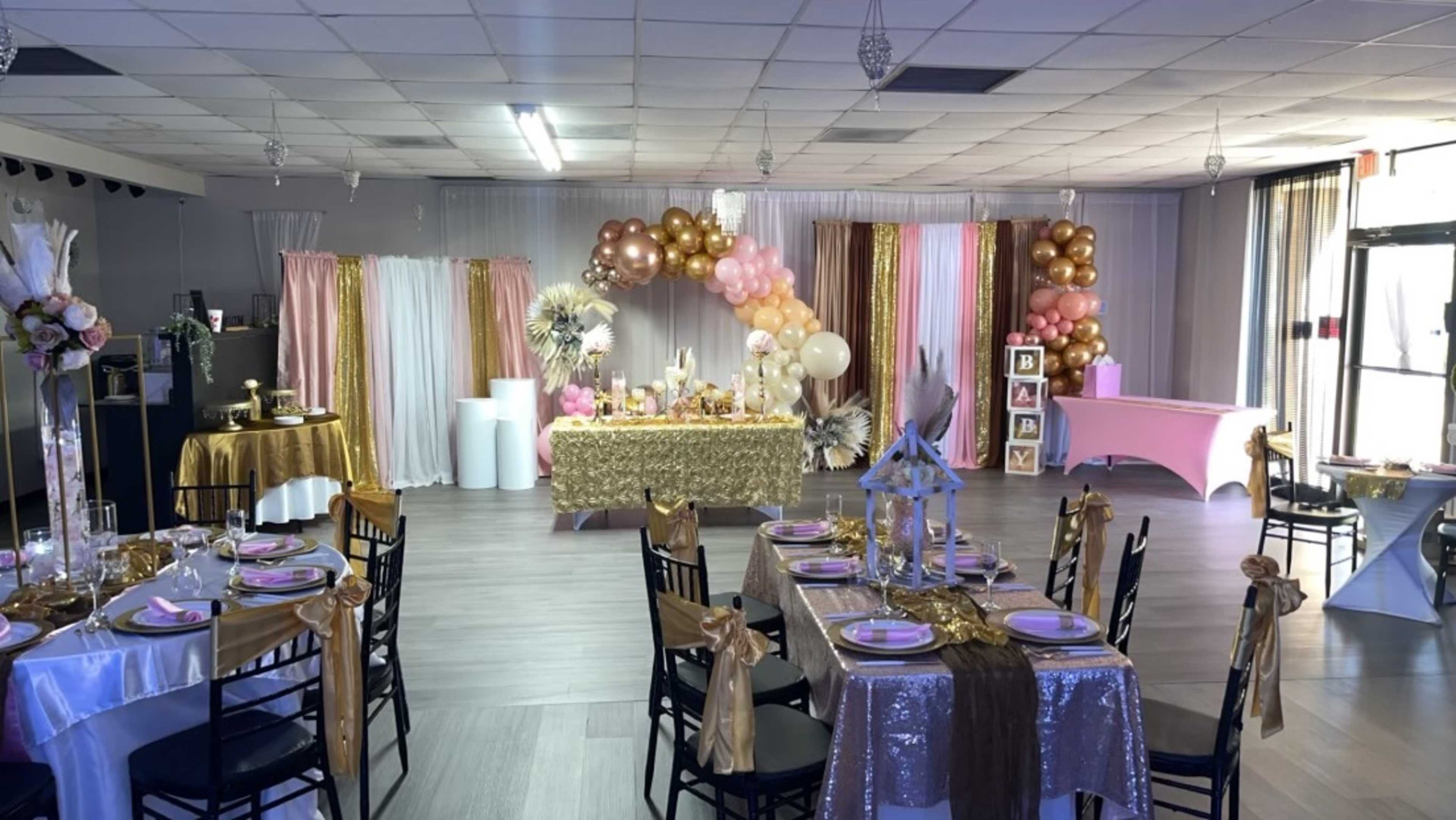 A decorated event space with tables set for a gathering, featuring gold and pink décor, balloons, and a dessert table.