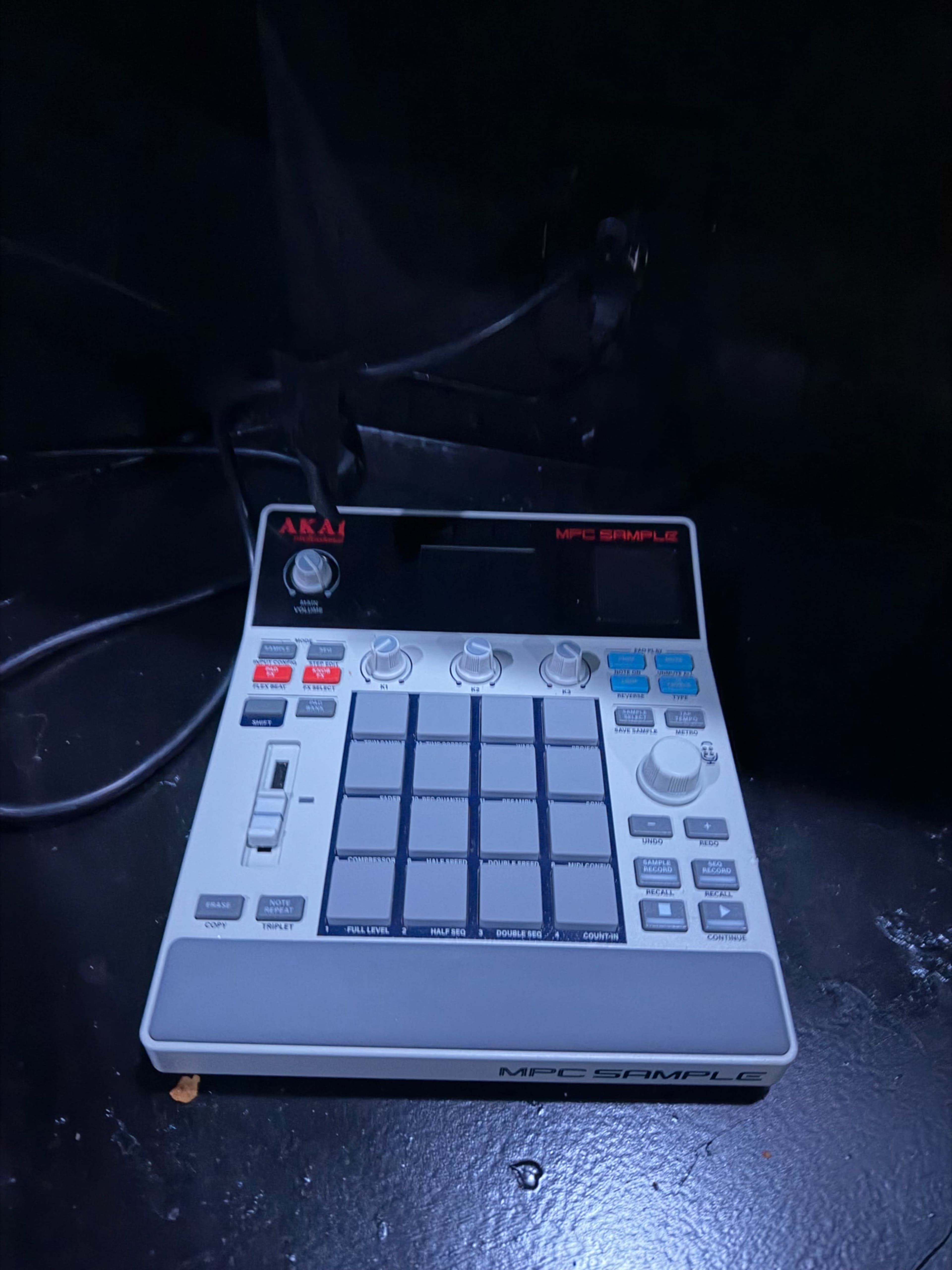 The image shows an Akai MPC Sampler with a grid of pads and various controls on its surface.