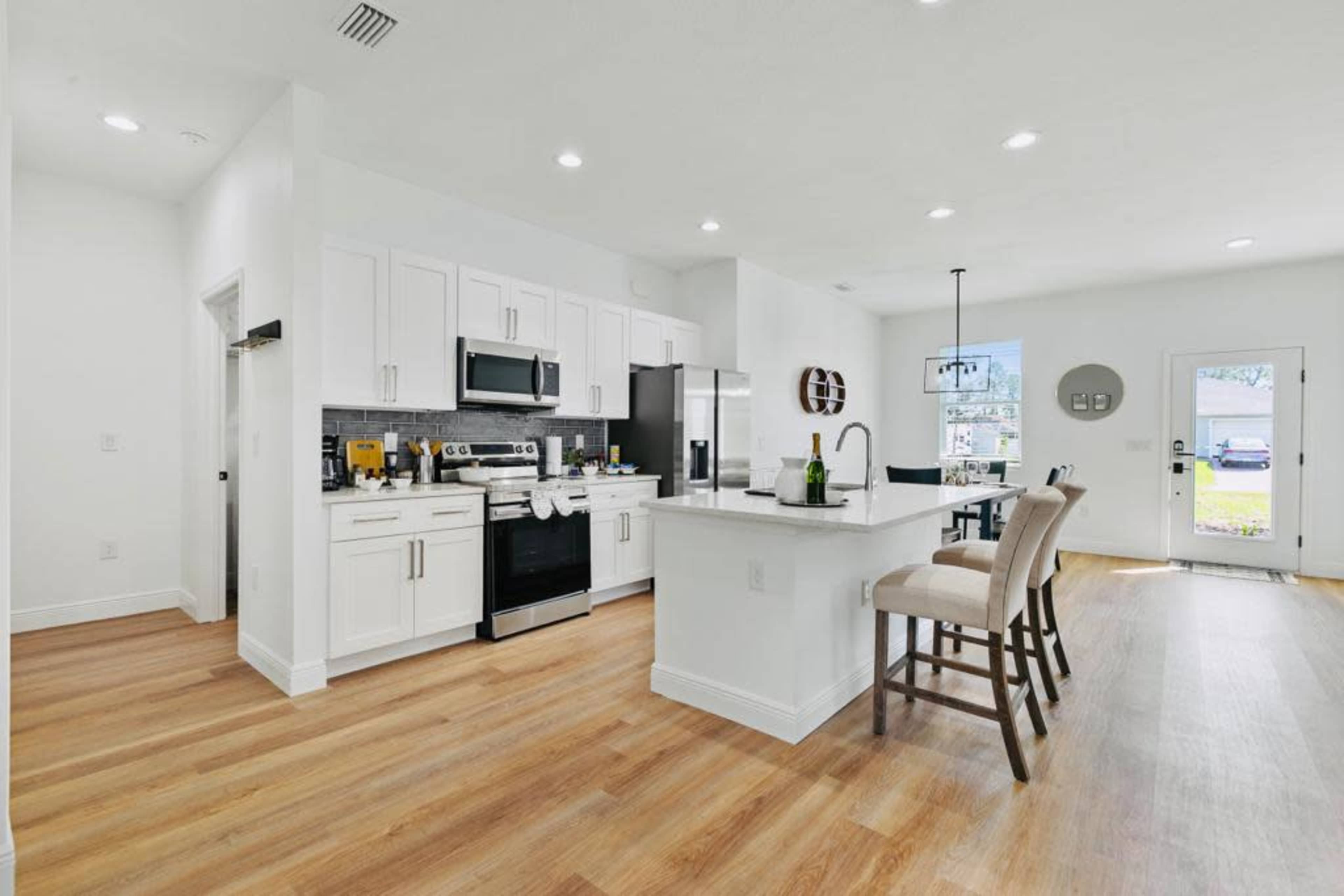 The image shows a modern kitchen with white cabinetry, stainless steel appliances, and a breakfast bar with three stools, all set in a spacious, well-lit area with hardwood flooring.
