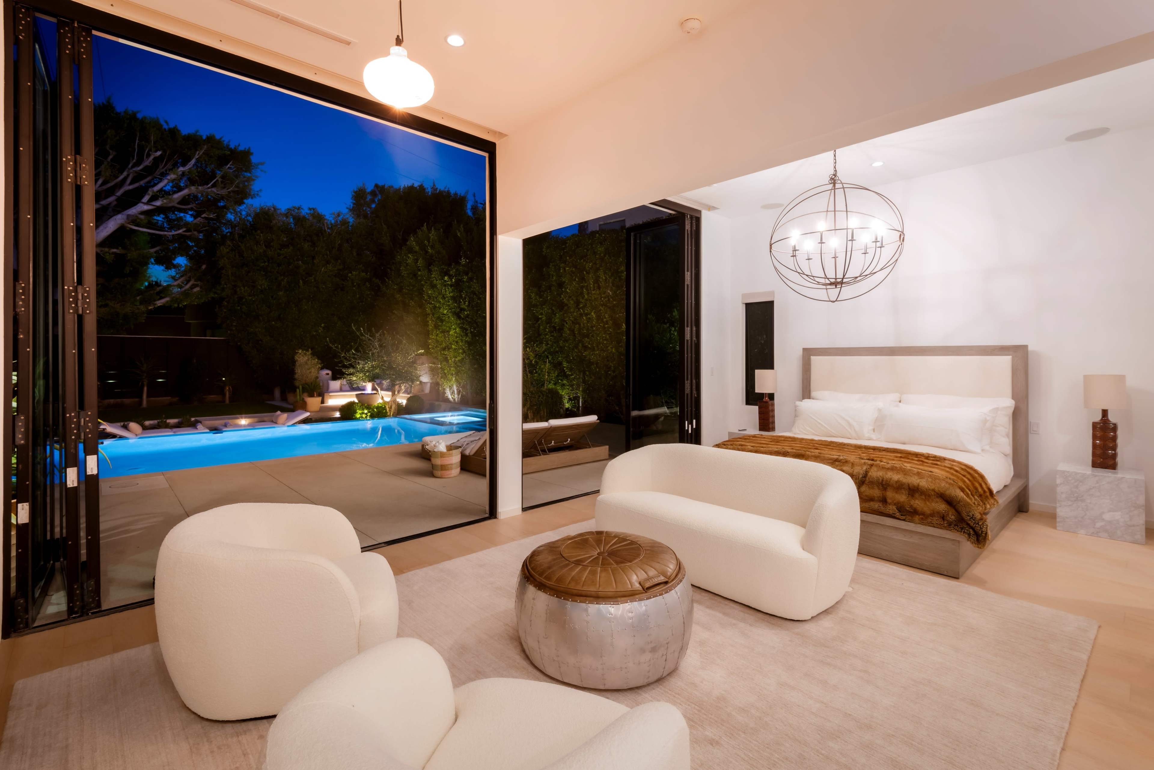 The image shows a modern bedroom that opens to an outdoor pool area, featuring large glass doors and minimalistic furniture.