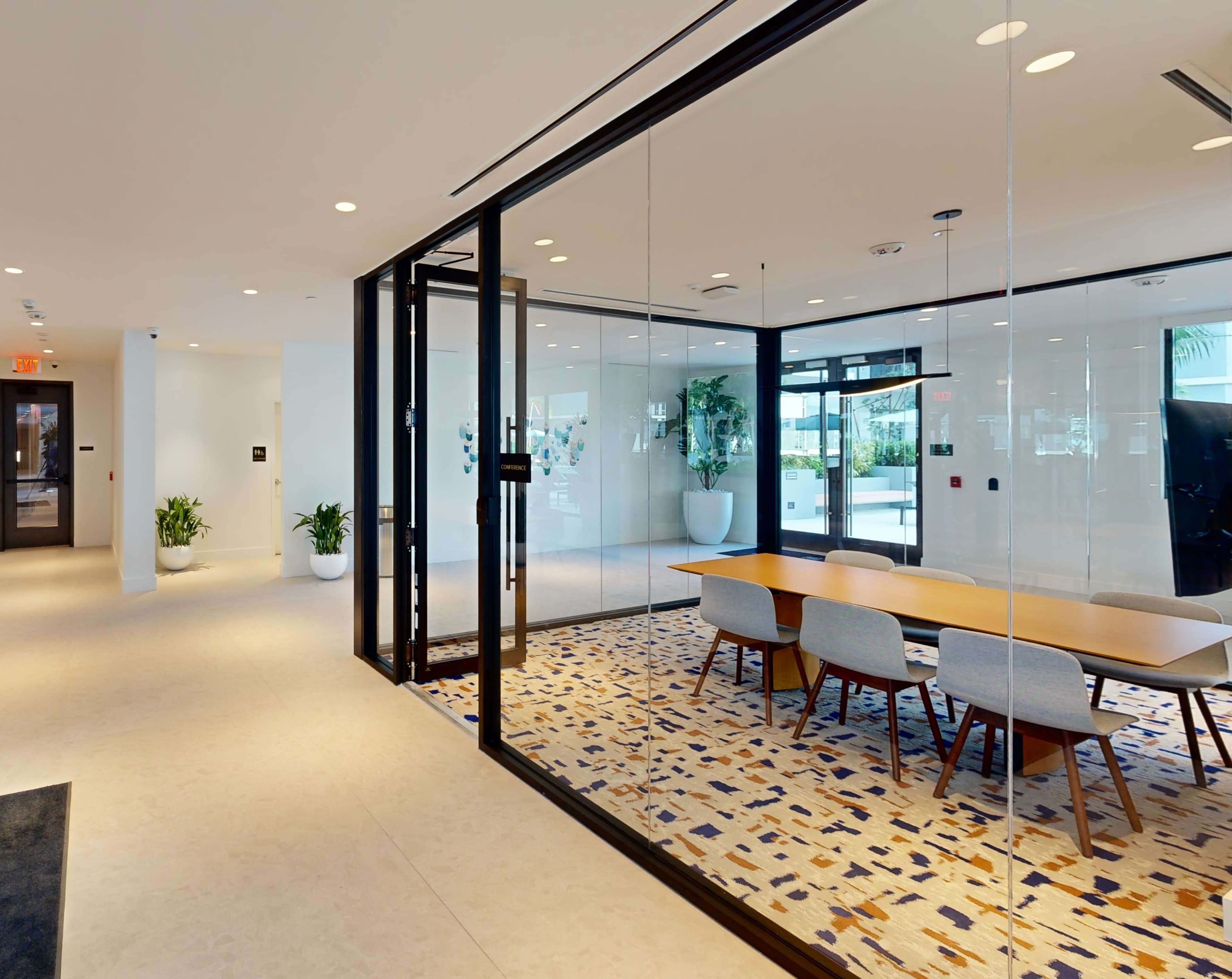 The image shows a modern office space featuring a glass-walled conference room with a large wooden table and several chairs, adjacent to an open area with potted plants.