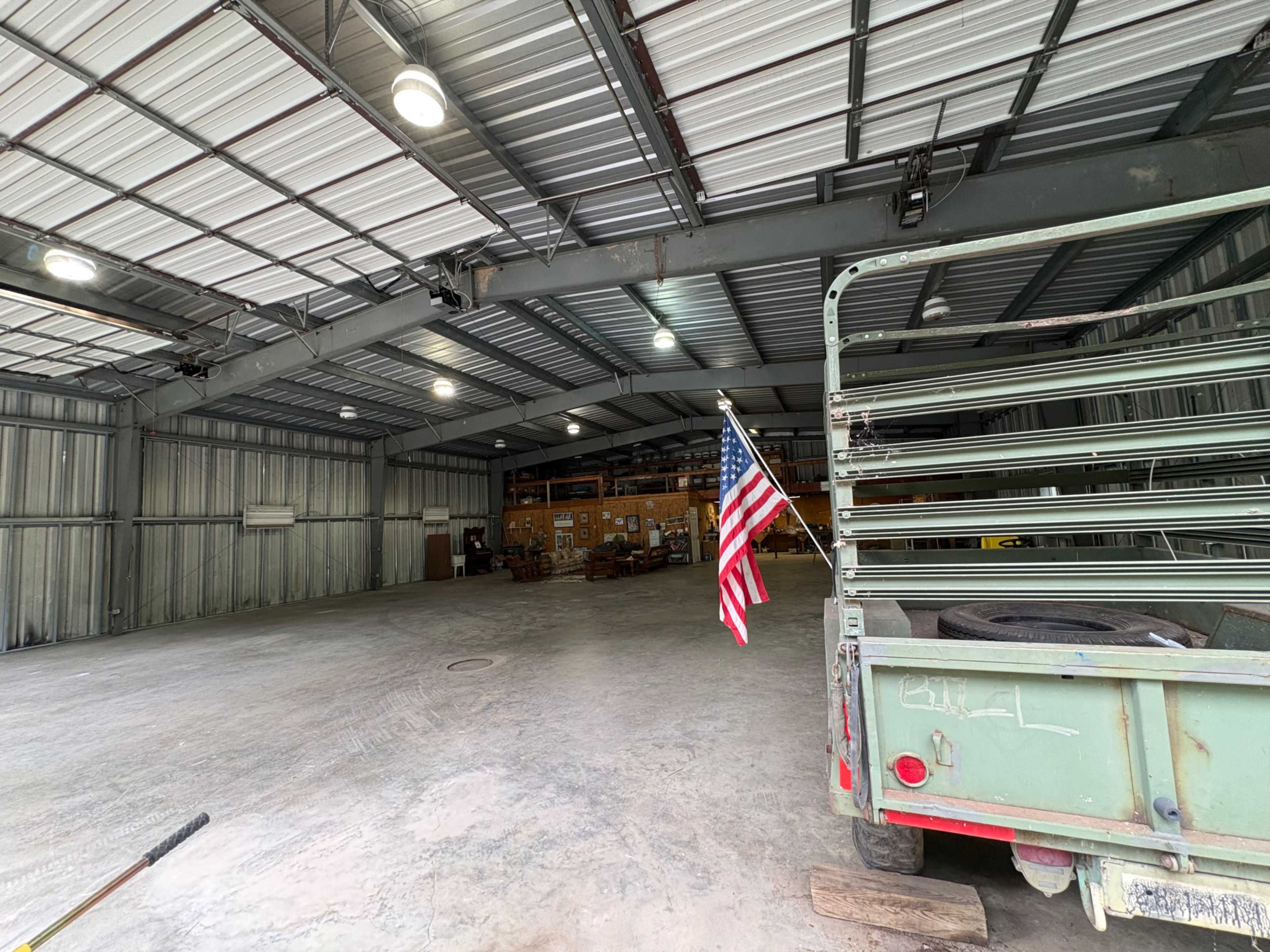 The image shows the interior of a large warehouse with metal siding, a concrete floor, and a parked trailer displaying an American flag near the entrance.