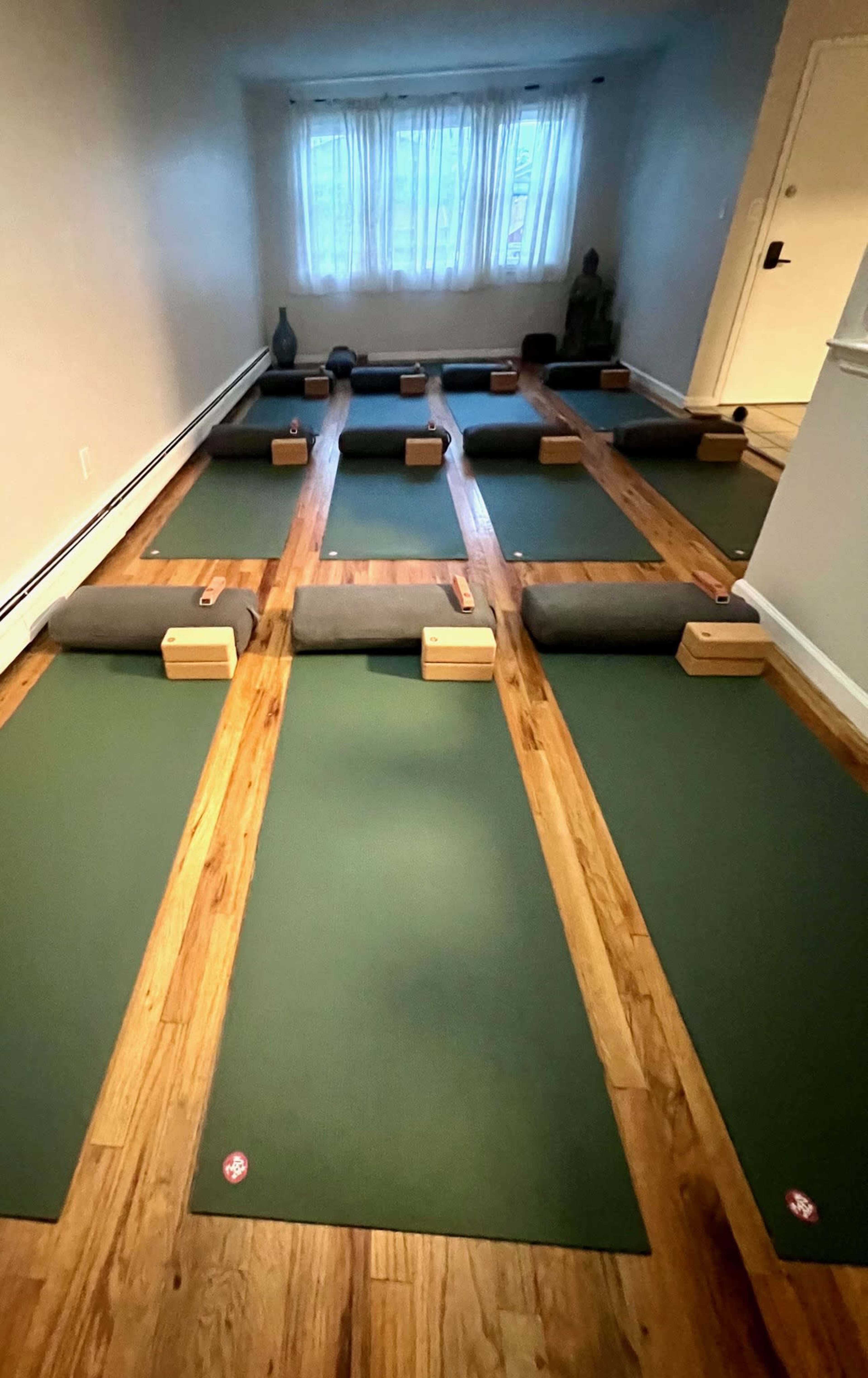 The image shows a yoga studio with neatly arranged green mats and props, including blocks and bolsters, set up on a wooden floor under soft lighting.