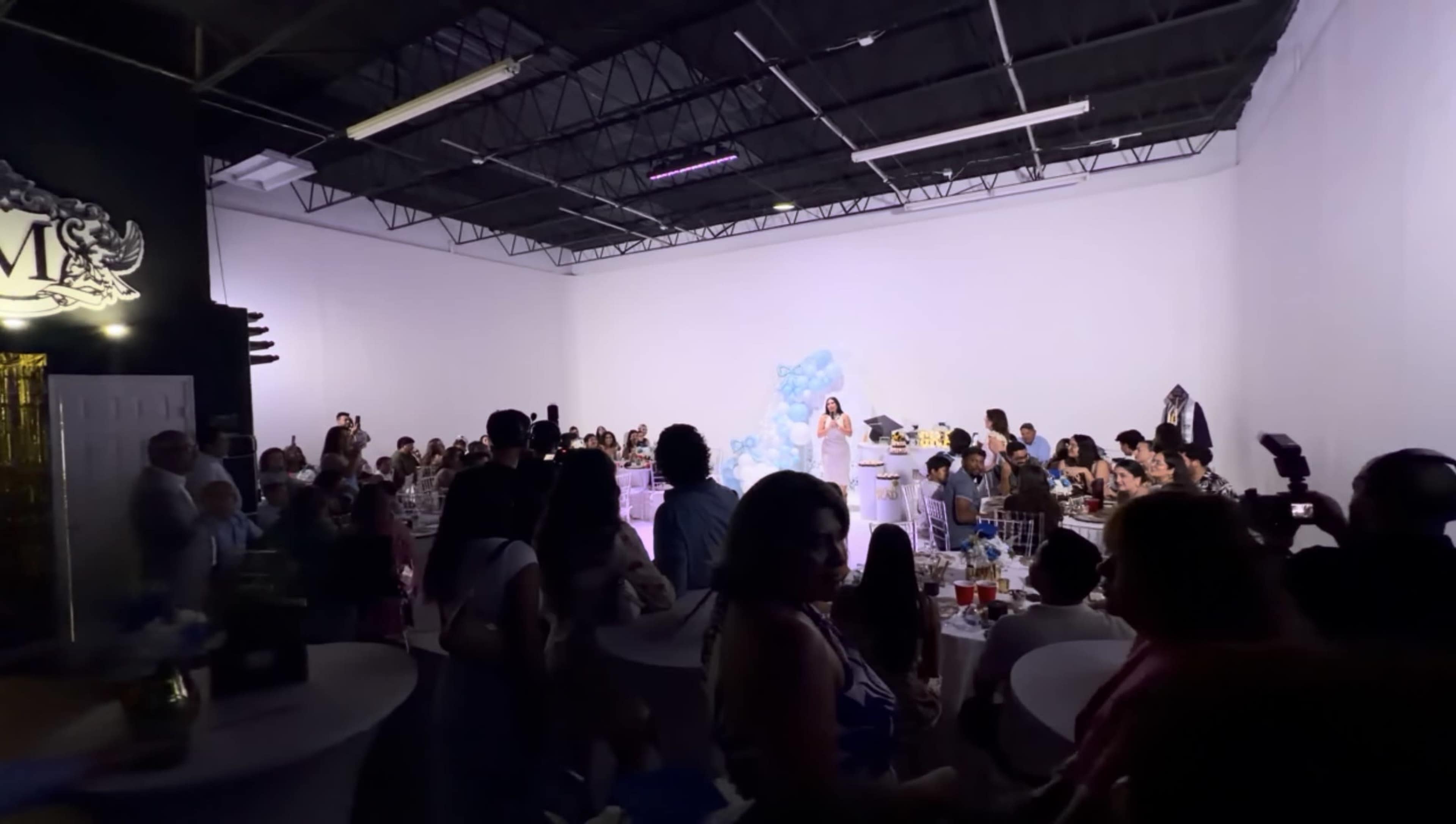A crowd gathers in a spacious event venue for a celebration, with tables arranged and a person speaking in front of a decorative backdrop.
