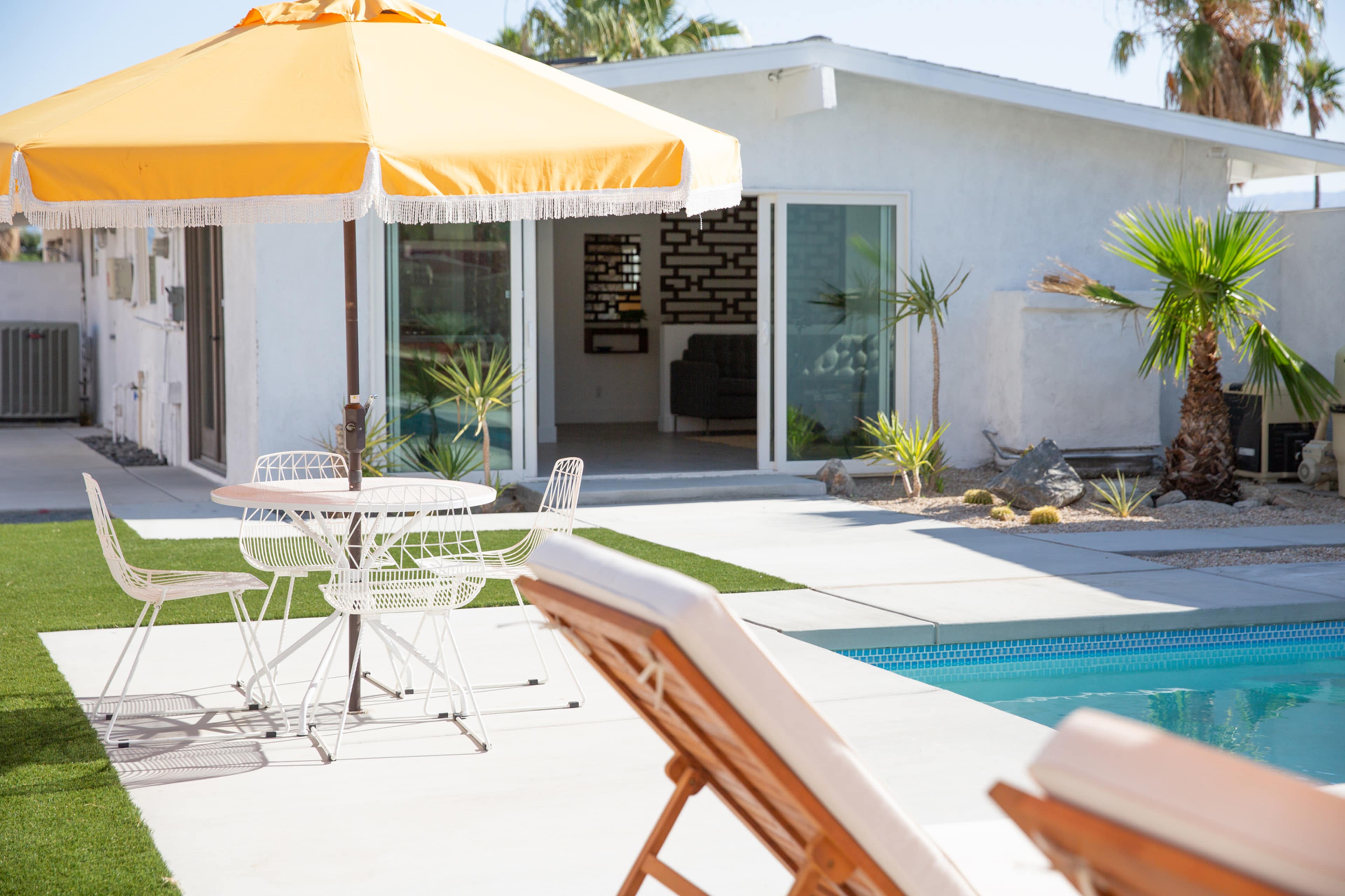 A yellow umbrella shades a white patio table and chairs near a swimming pool, with a modern home and palm trees in the background.