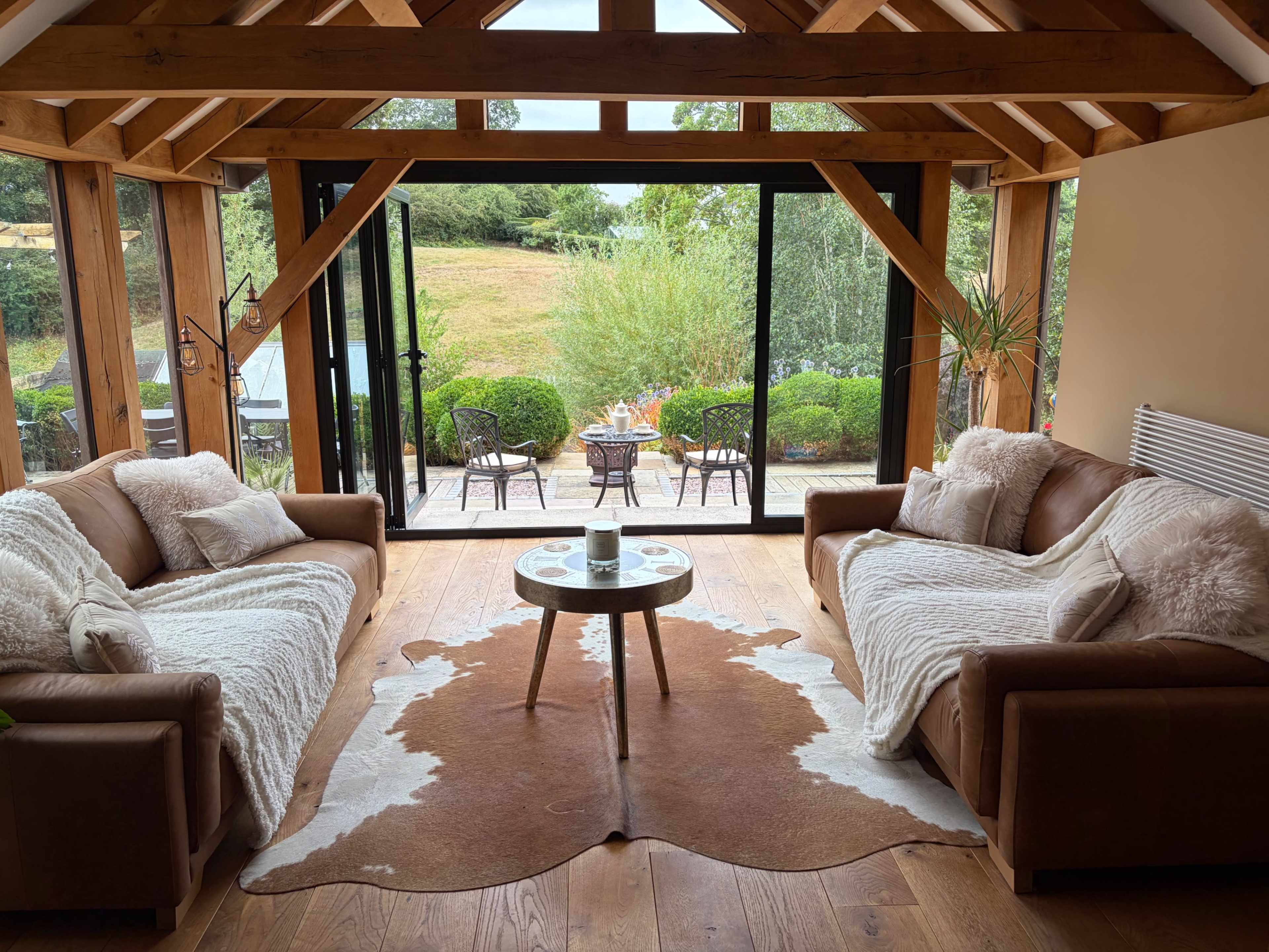 A cozy living room features two brown sofas with white throws, a circular coffee table on a cowhide rug, and large glass doors opening to a serene outdoor view.