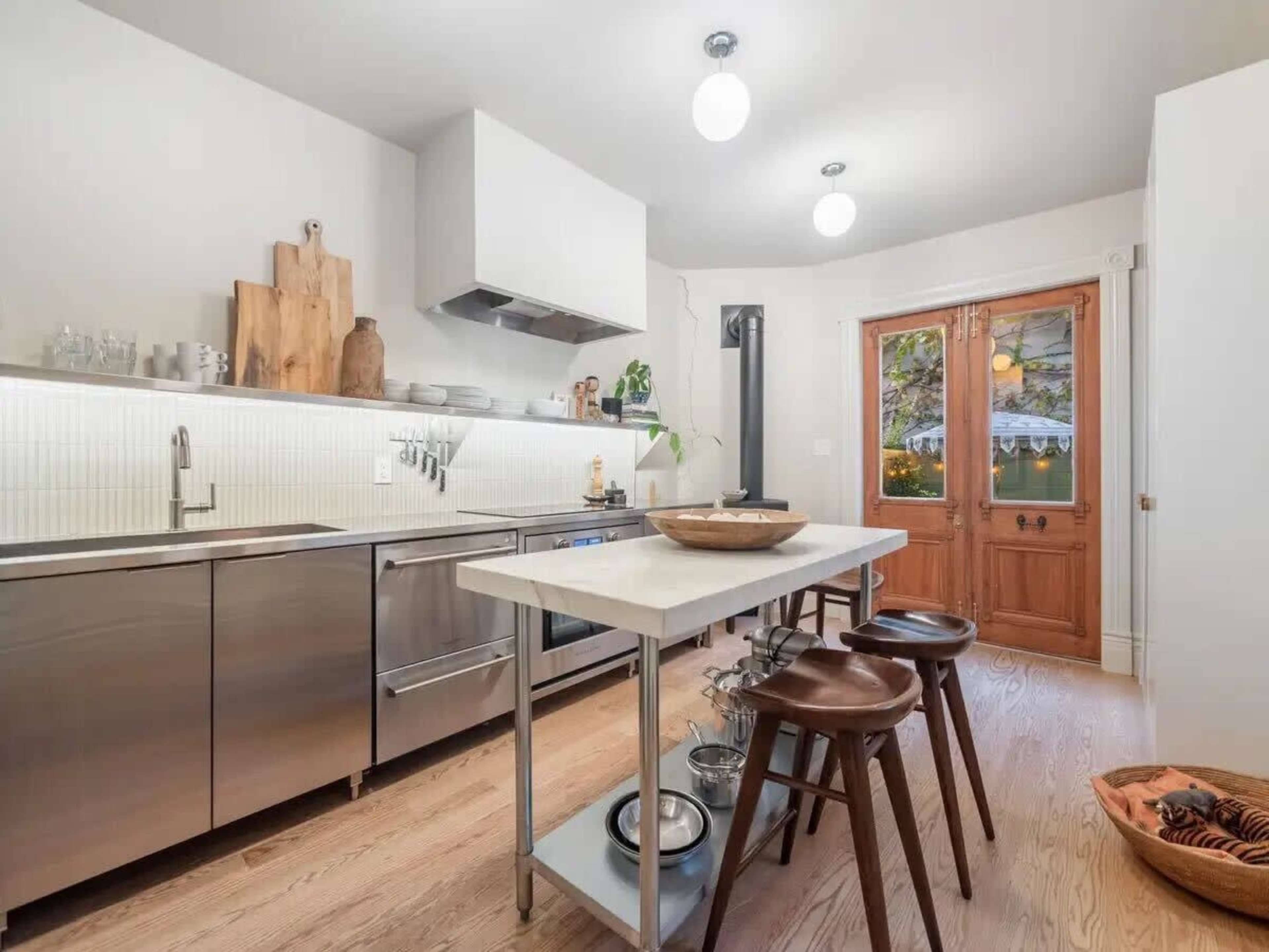 The image shows a modern kitchen featuring stainless steel appliances, a central island with a marble top, wooden bar stools, and a door leading outside.
