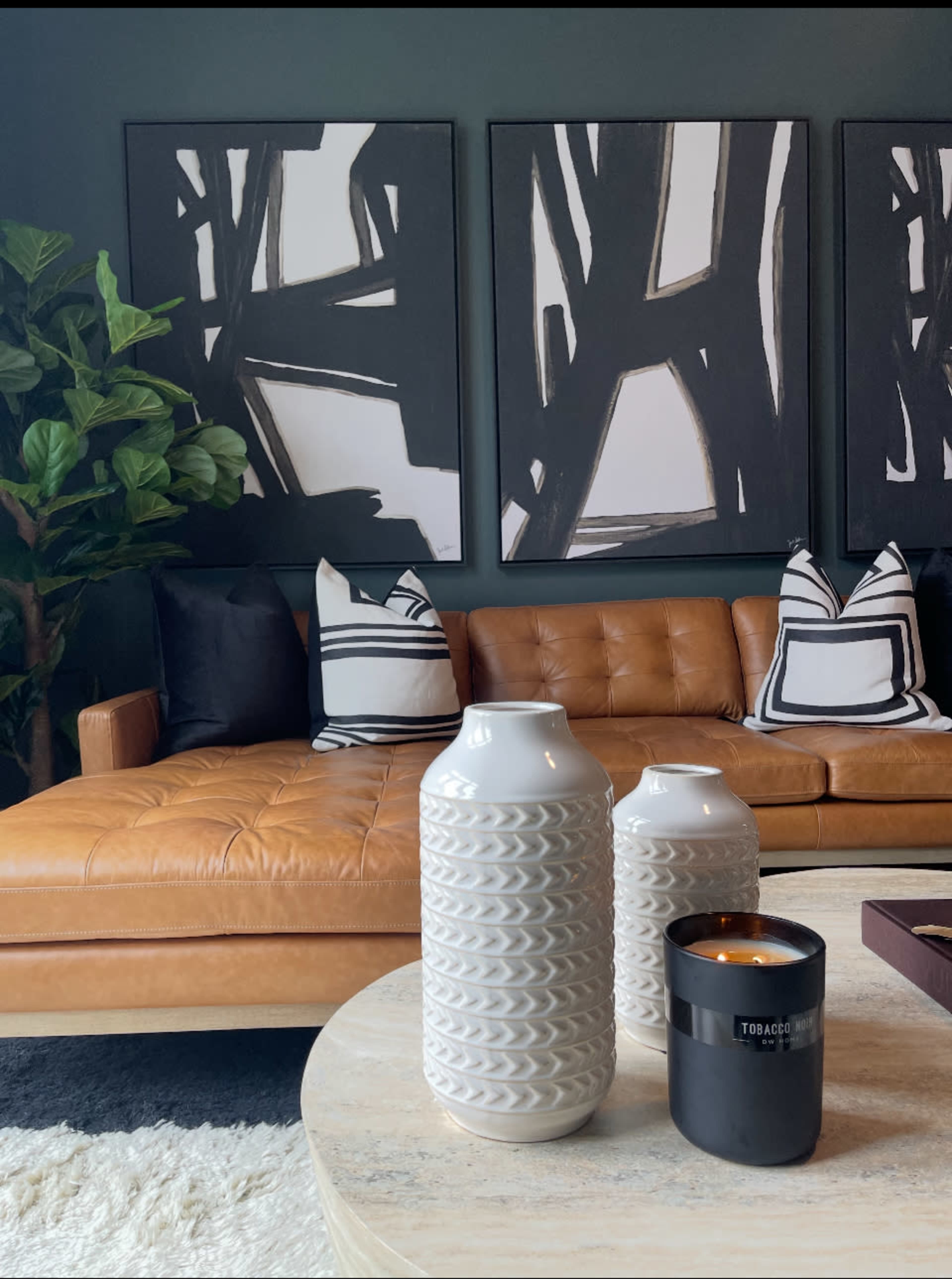 The image shows a stylish living room featuring a brown leather sofa adorned with black and white pillows, two patterned vases, and a candle on a round coffee table, with abstract artwork hanging on the wall behind.
