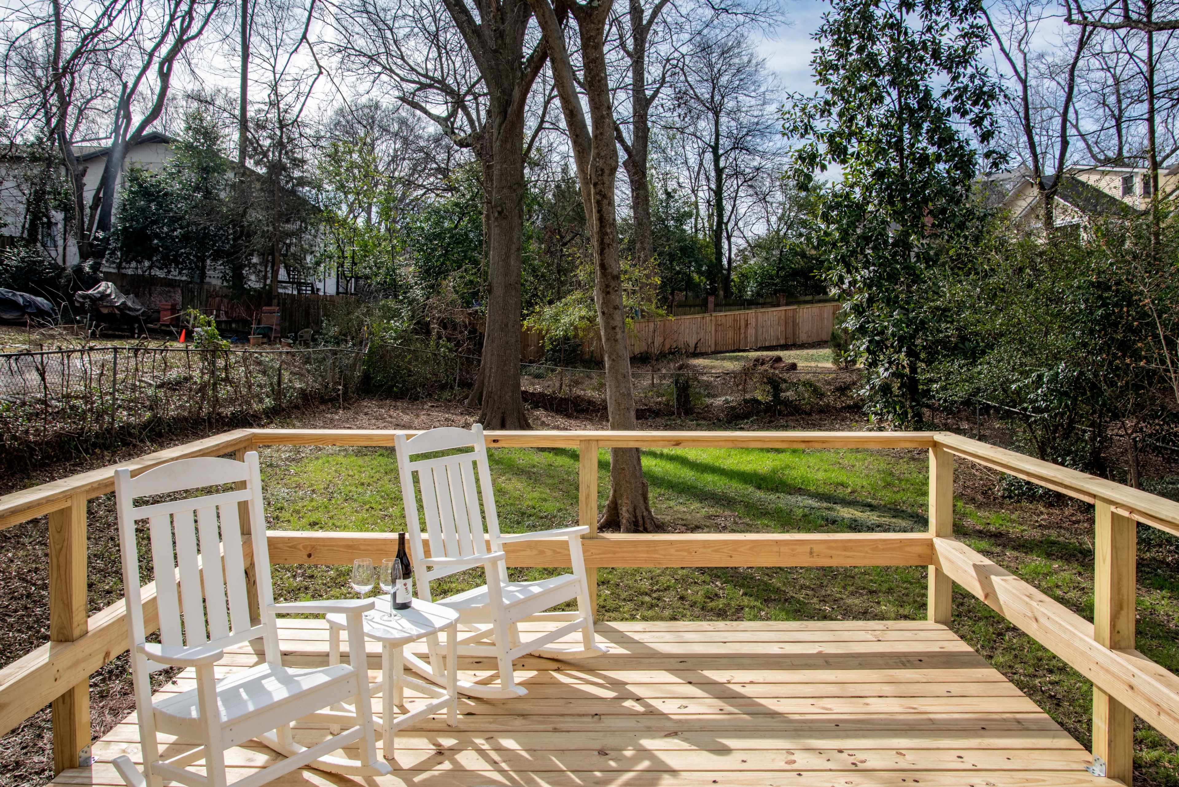 A wooden deck features two white rocking chairs and a bottle with glasses, overlooking a grassy yard surrounded by trees.
