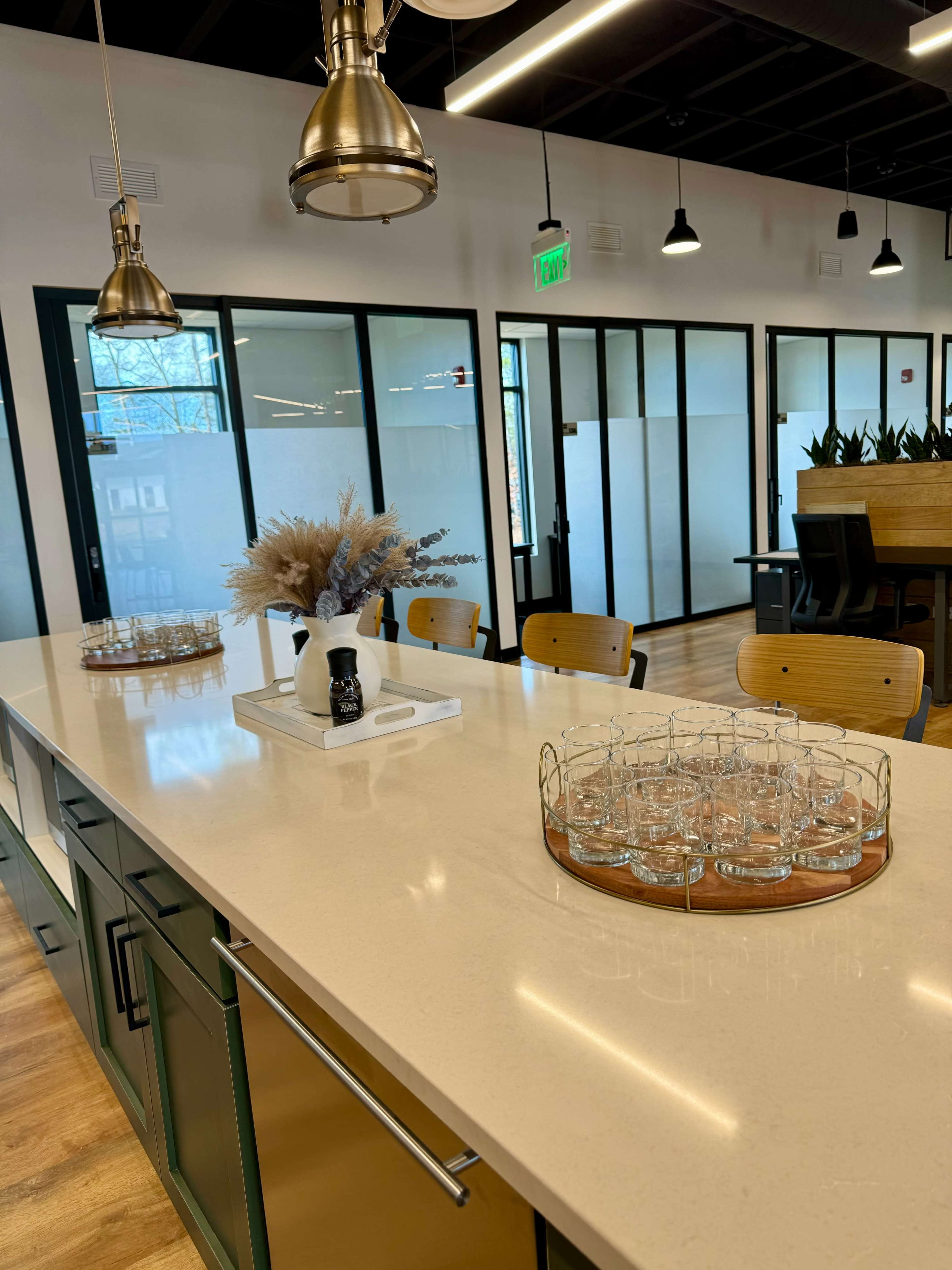 The image shows a modern office kitchen area featuring a countertop with glassware neatly arranged on a wooden tray and decorative plants in the background.