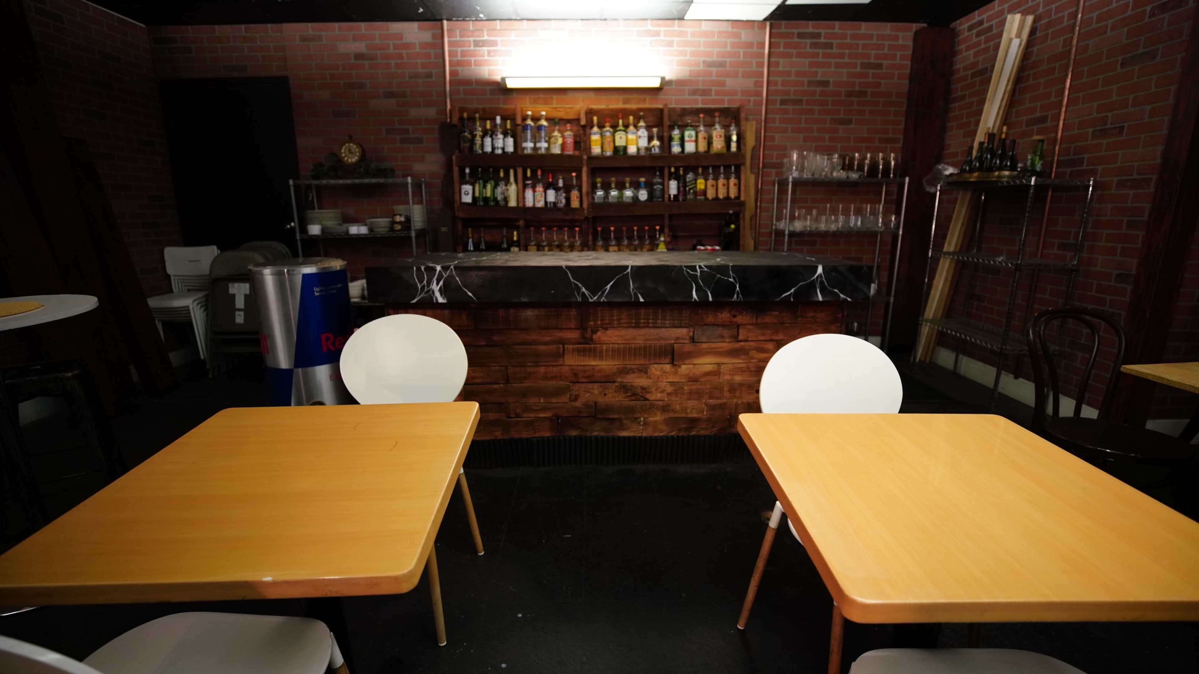 A bar area with a wooden counter and shelves stocked with various bottles, set in a room featuring brick walls and simple wooden tables.