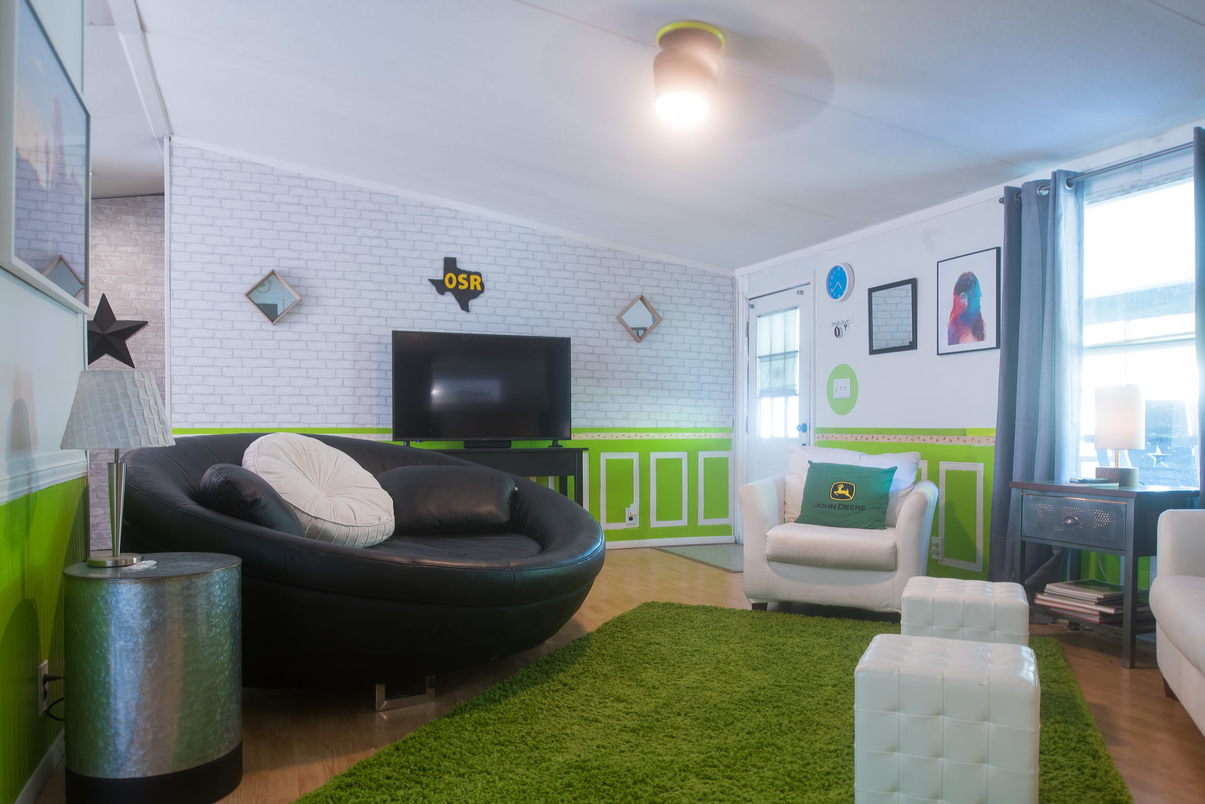 The room features a modern design with a black circular sofa, a white armchair, a green rug, and a wall-mounted television surrounded by vibrant green and white accents.