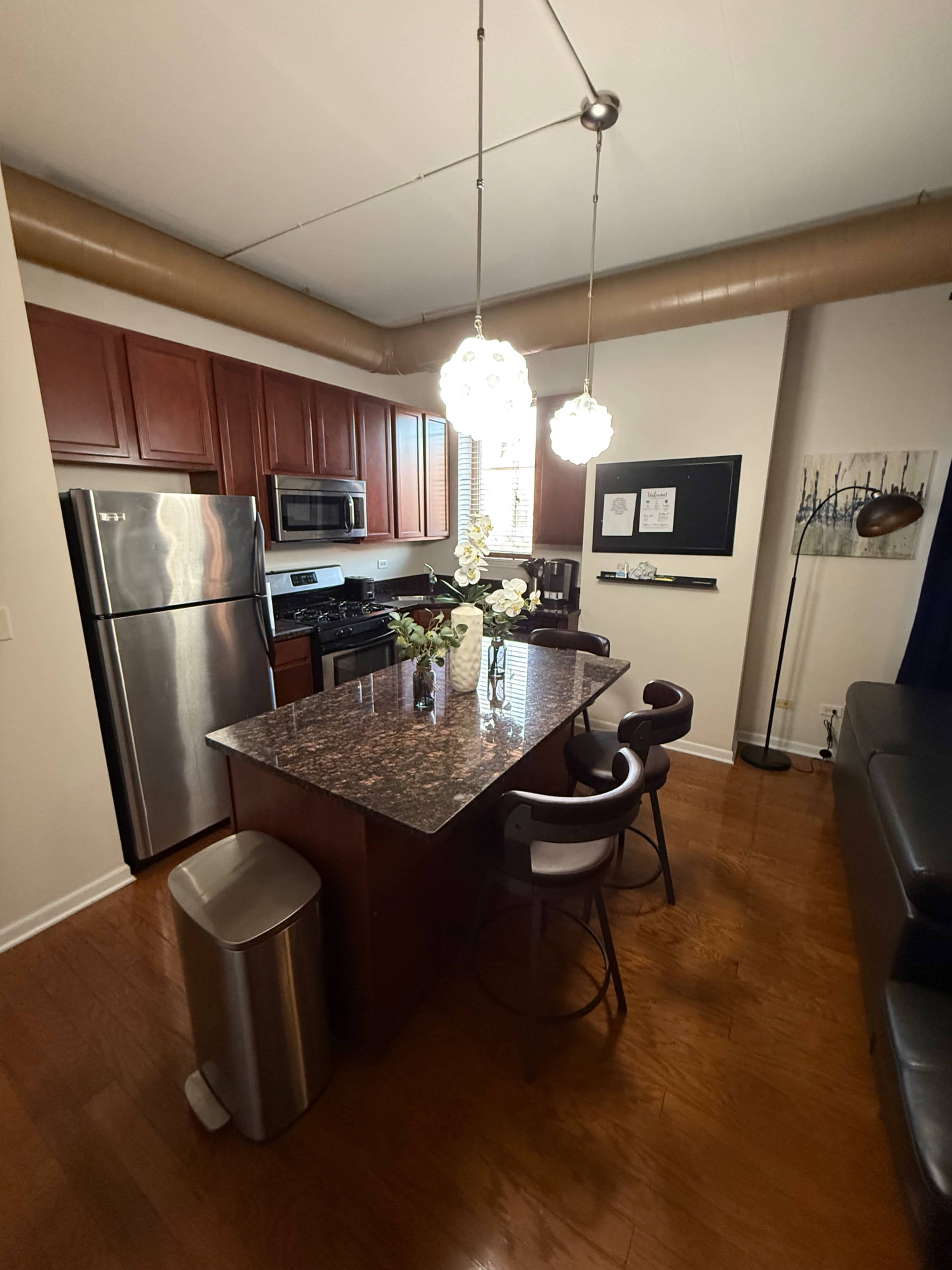 The image shows a modern kitchen featuring stainless steel appliances, dark wood cabinets, a granite countertop island with bar stools, and decorative lighting.